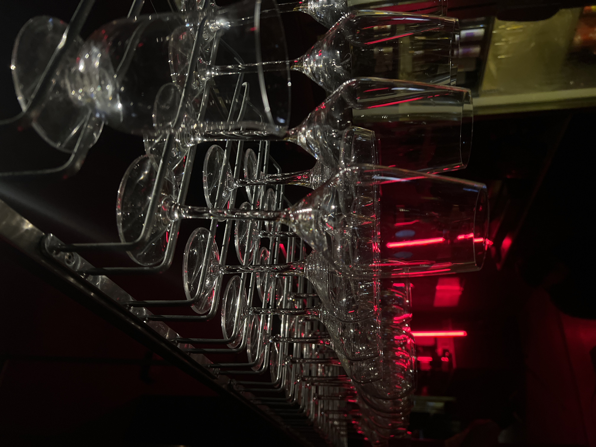 The image depicts rows of clean wine glasses hanging upside down in a metal rack. The background is dimly lit, with red lighting in the distance. The location, Mengwi, Indonesia, is not readily apparent from the visual content of the image. There are no people visible.