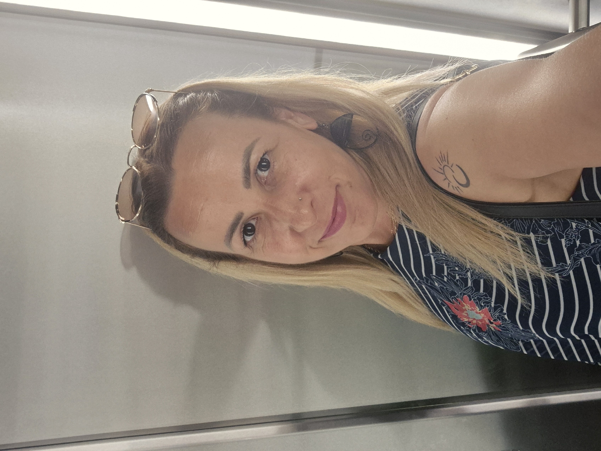 A woman with blonde hair and sunglasses on her head is shown from the shoulders up, standing in front of a neutral gray wall. She is wearing a dark blue and white striped shirt with a floral pattern on the chest. She has a small nose ring and a tattoo of a crescent moon with a sun on her right shoulder. She is smiling at the camera. The lighting suggests an indoor setting, possibly an elevator or hallway.