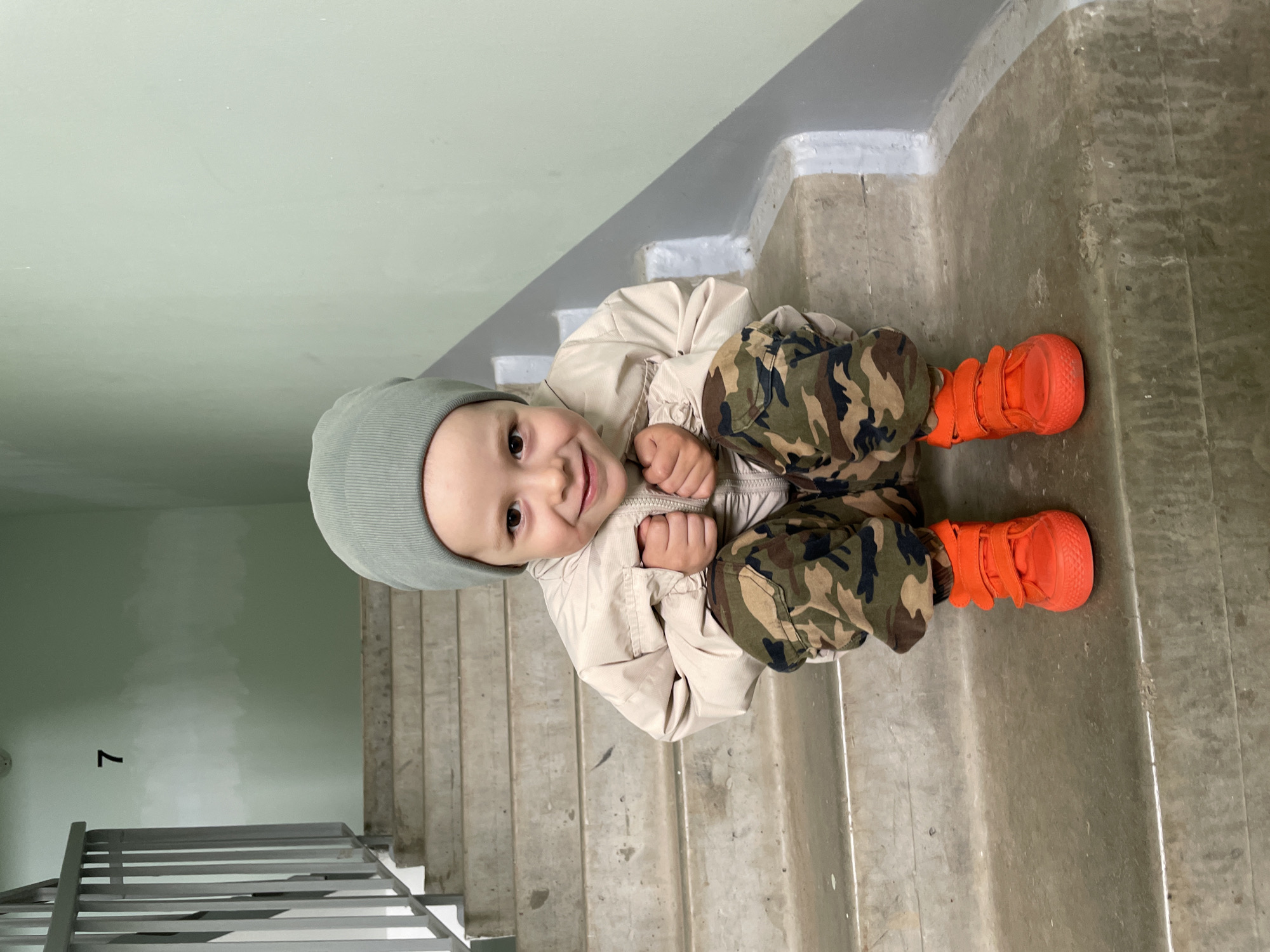 A young child, appearing to be a toddler or preschooler, sits on a concrete staircase in an indoor stairwell. The child is looking upwards and smiling directly at the camera, conveying a happy and playful emotion.
The child is dressed in a light green beanie, a light beige padded jacket, camouflage-patterned pants in shades of green, brown, and black, and bright, vibrant orange shoes with velcro straps. Their hands are clasped together in front of their chest.
The setting is a utilitarian stairwell with a concrete staircase that has a somewhat rough, weathered texture. The walls are painted a plain light green. A dark gray metal railing is visible on the left side of the frame, running alongside the stairs. In the distance, on the light green wall, a black number "7" is visible. The lighting suggests an indoor daytime scene.