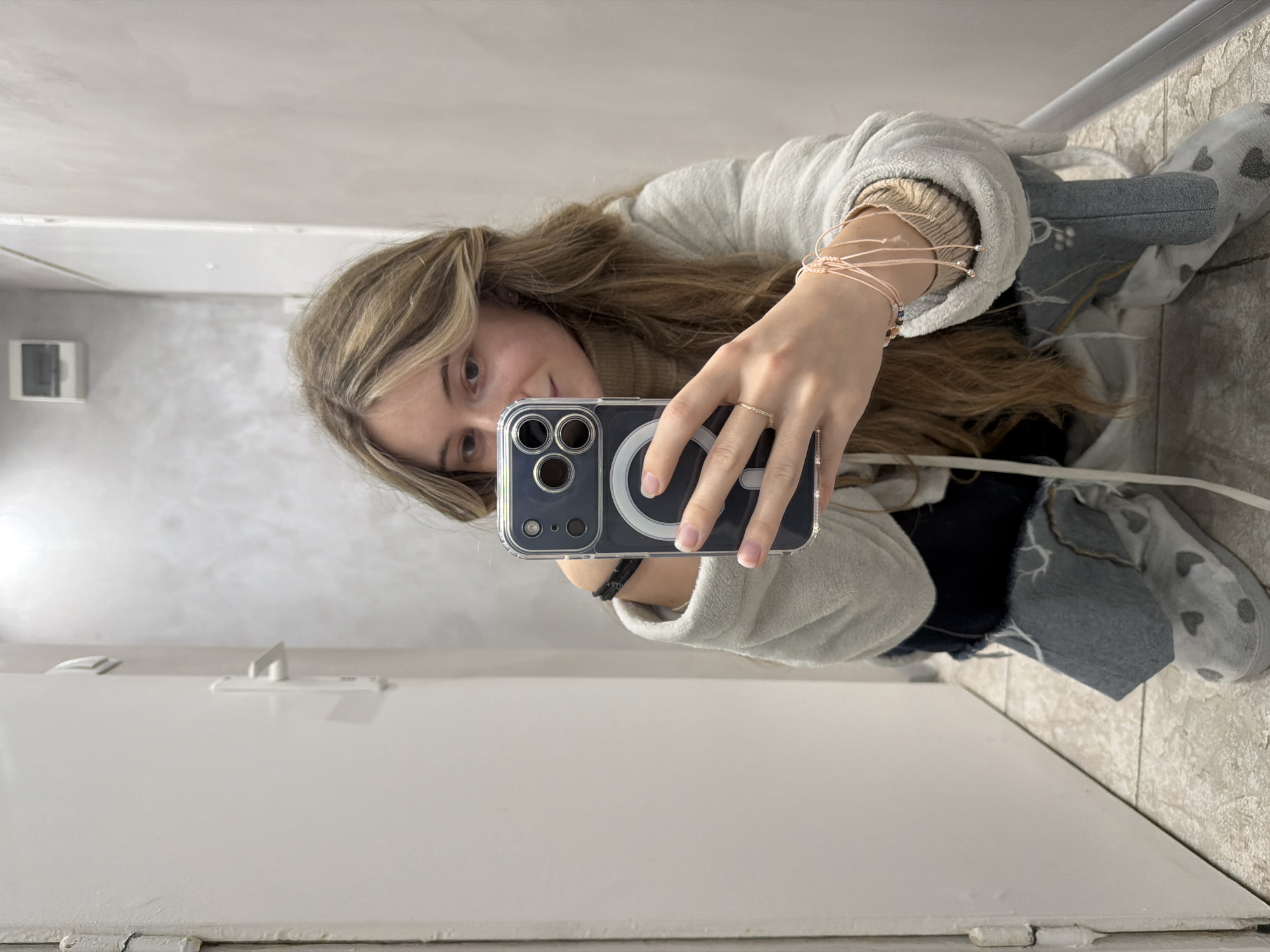 A woman is taking a selfie in what appears to be a bathroom. She is holding an iPhone with a clear case that reveals a MagSafe charger. She is wearing a light-colored robe, a tan turtleneck sweater, and multiple string bracelets on her wrist. The background includes a white door, tiled walls, and tile flooring. There is a small electrical panel on the wall. The location cannot be identified as Plovdiv, Bulgaria, or any specific place based on the provided visual information.