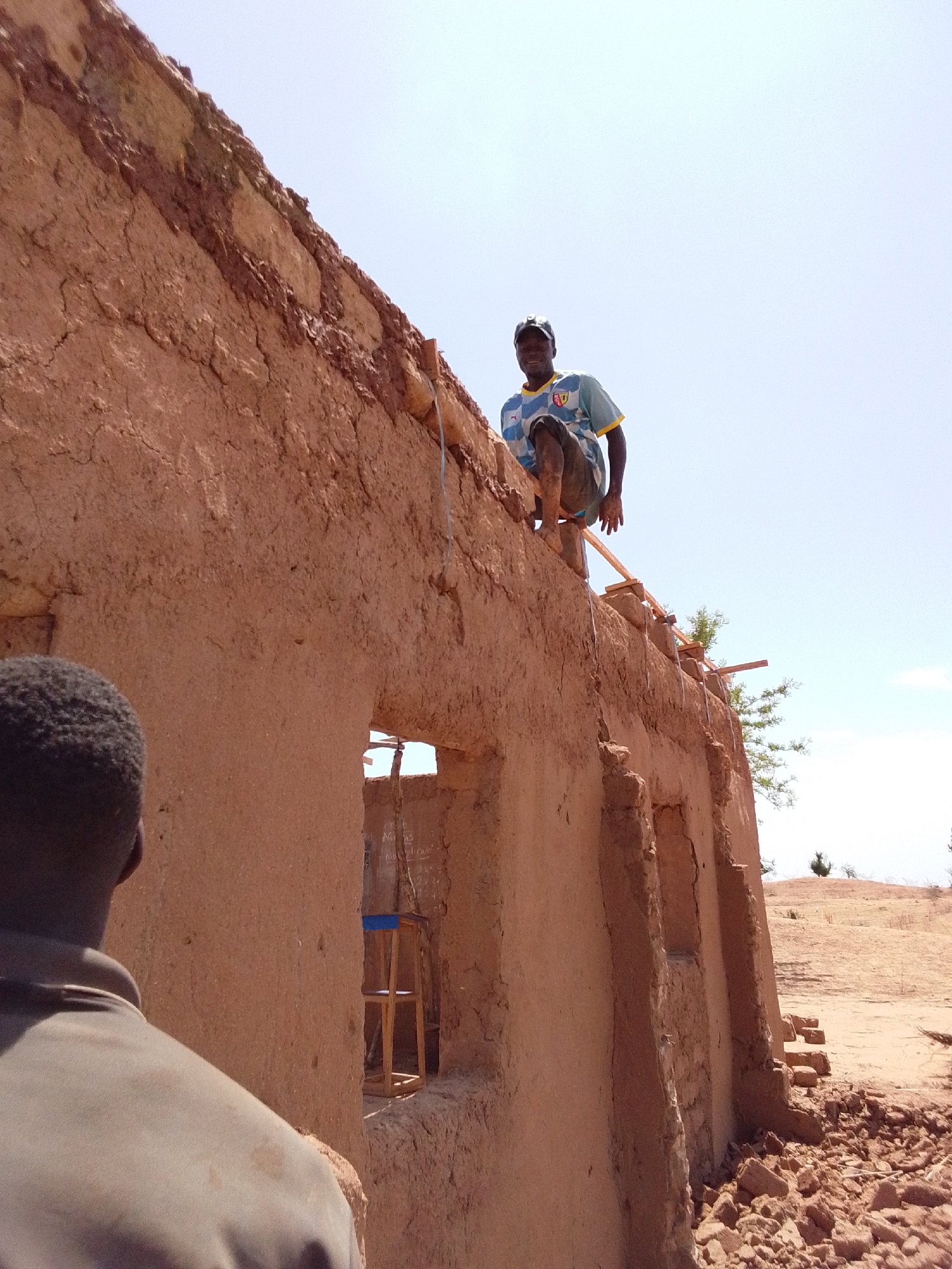 The image shows a man in a blue and yellow jersey standing on the partially constructed roof of a mud building in Mulke Barikin Sayejo, Nigeria. Another man, seen from the back, is standing at ground level near the building. The building, made of mud, has visible window openings, and a chair is seen inside one of the openings. Construction materials are visible on the roof and debris from the building lies on the ground. The landscape behind the building appears dry and hilly under a clear sky.