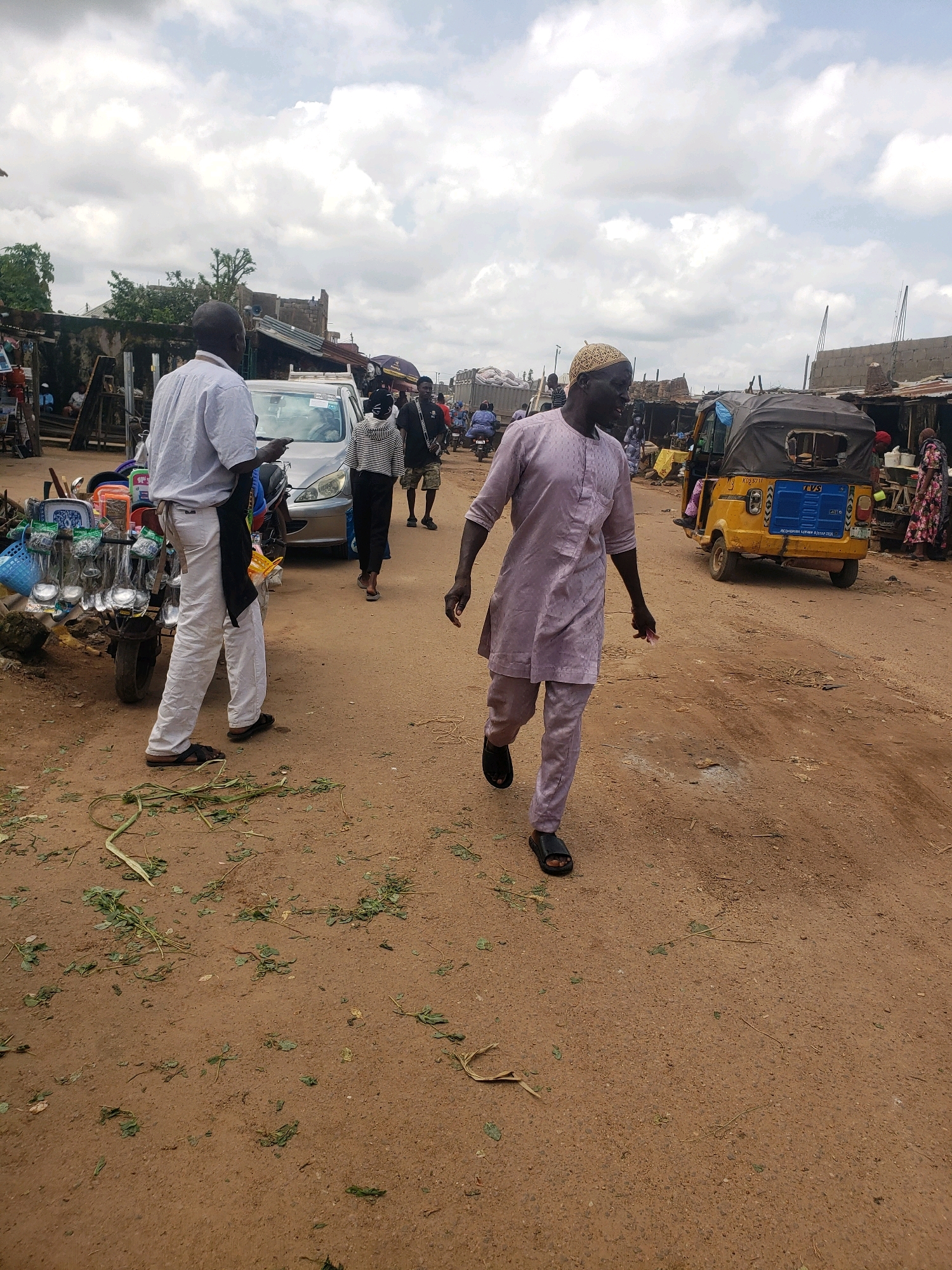 A street scene in Sabon Tasha, Nigeria, features a bustling outdoor market environment under a cloudy sky. In the foreground, a man in a light purple traditional outfit with matching pants and a patterned shirt walks from left to right across a dirt road. He is wearing black sandals and has short, bleached hair. To his left, another man wearing a white shirt and white pants, with a black apron tied around his waist, stands beside a cart laden with various kitchen utensils, including ladles, strainers, and bowls. Behind him, a silver car is parked. Further in the background, a yellow auto-rickshaw is visible, along with several market stalls and other pedestrians. The ground is a mixture of dirt and scattered green leaves.