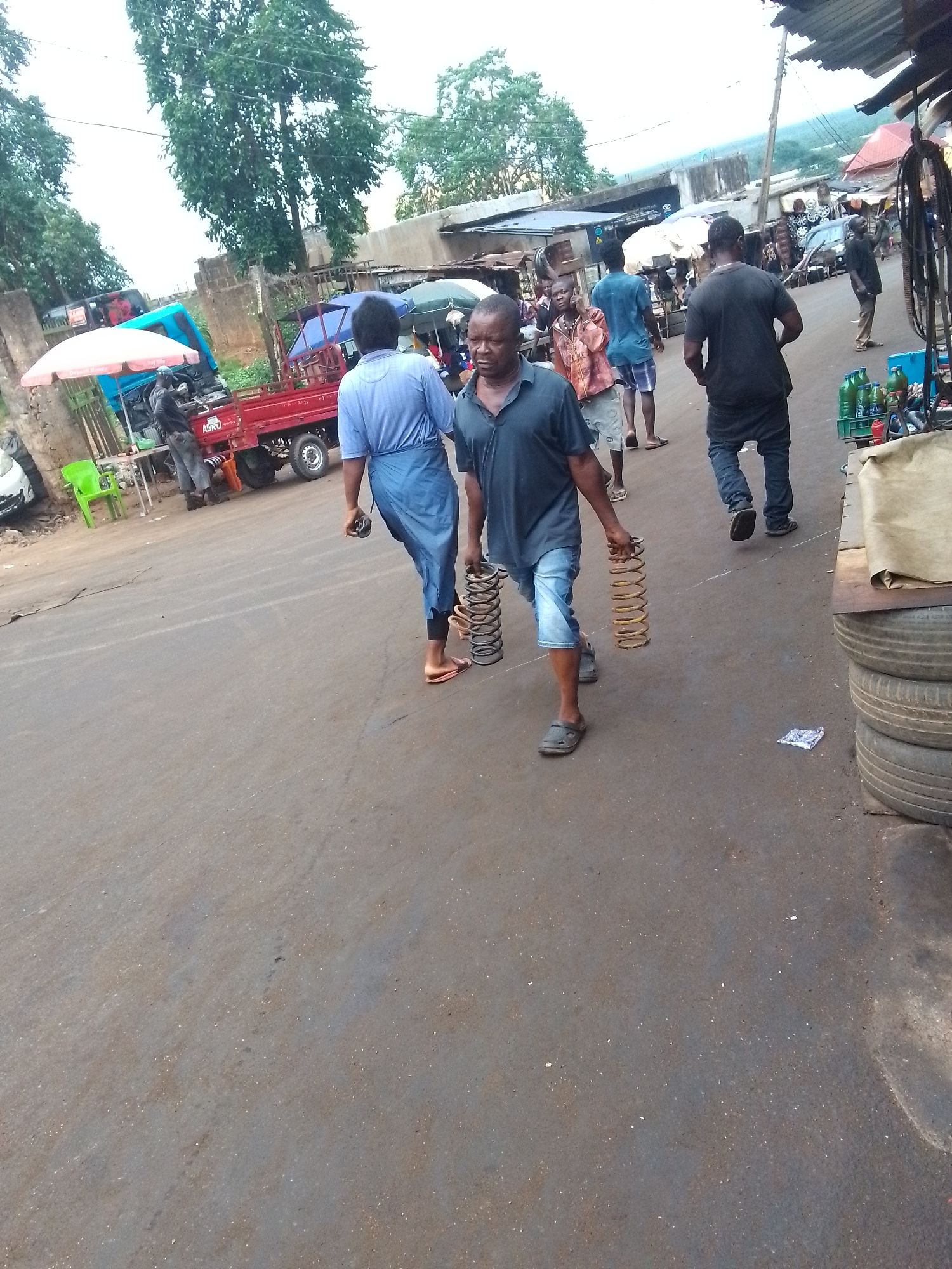 The image shows an outdoor street scene in Onitsha, Nigeria. The street is unpaved. A man in a dark blue shirt and light blue shorts walks along the street, carrying two large metal springs, one in each hand. A woman in a light blue blouse and a dark blue wrap follows behind him. Several other people are walking on the street. In the background, there is a red truck, a blue truck, and a green umbrella. A stack of tires is visible in the lower right corner of the image.