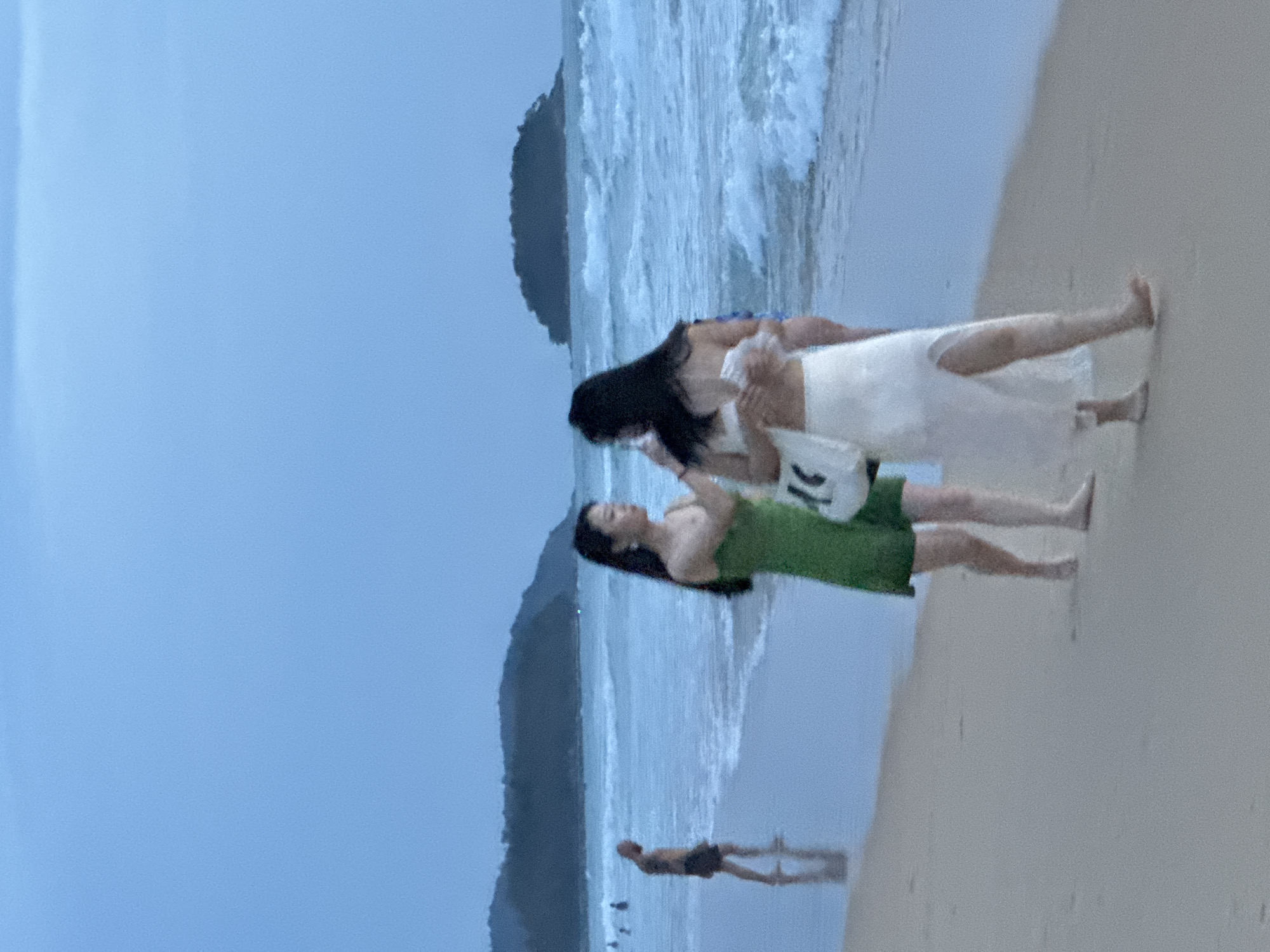 At dusk on Karon Beach in Karon, Thailand, a man with a shaved head and wearing dark shorts stands on the wet sand near the waterline, facing away from the camera. Two women are walking on the beach, closer to the foreground. The woman on the left wears a short green dress and has dark hair. The woman on the right wears a white top and a long white skirt with a slit, and also has dark hair. She is carrying a white bag. The ocean is visible behind them with waves crashing on the shore. In the distance, a dark, tree-covered island or peninsula is silhouetted against the hazy sky. Several other figures are visible in the water further down the beach. The overall lighting suggests late evening or early morning, with soft, diffused light.