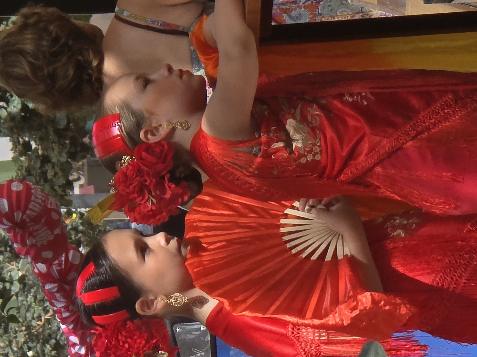 Two young girls, dressed in traditional Spanish flamenco attire, are pictured in a close-up shot. The girl on the left wears a red dress with a fringe and a red hair accessory, holding a red fan. The girl on the right is also in red attire, with a red flower in her hair. Behind them, a woman with brown curly hair is visible, wearing a patterned strap top. The scene appears to be outdoors, possibly during a street festival, given the decorative red and white banner overhead, which has text that could suggest a Spanish locality. The background includes foliage and what appears to be a stall or display. The lighting suggests a sunny day. The image captures a moment of cultural celebration, likely during the Málaga Fair (Feria de Málaga), an annual event held in August that celebrates the city's Reconquista.