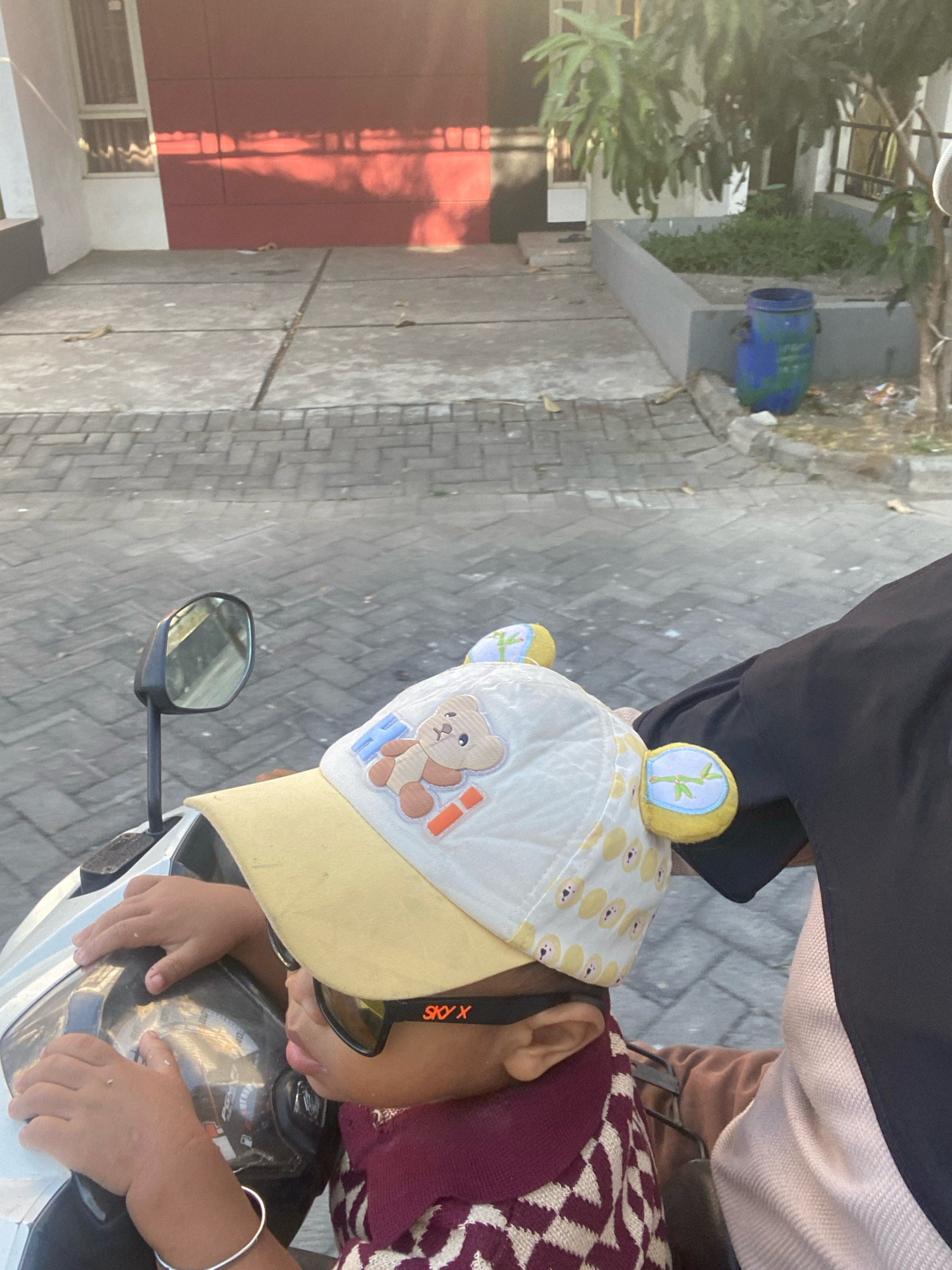 A young child, wearing sunglasses and a cap with bear ears, sits on a scooter in Gisik Cemandi, Indonesia. The child's hands are on the scooter's handlebars. The background shows a paved area in front of a building with a red wall and some trees. A blue trash bin is visible to the right. The scene is illuminated by sunlight.