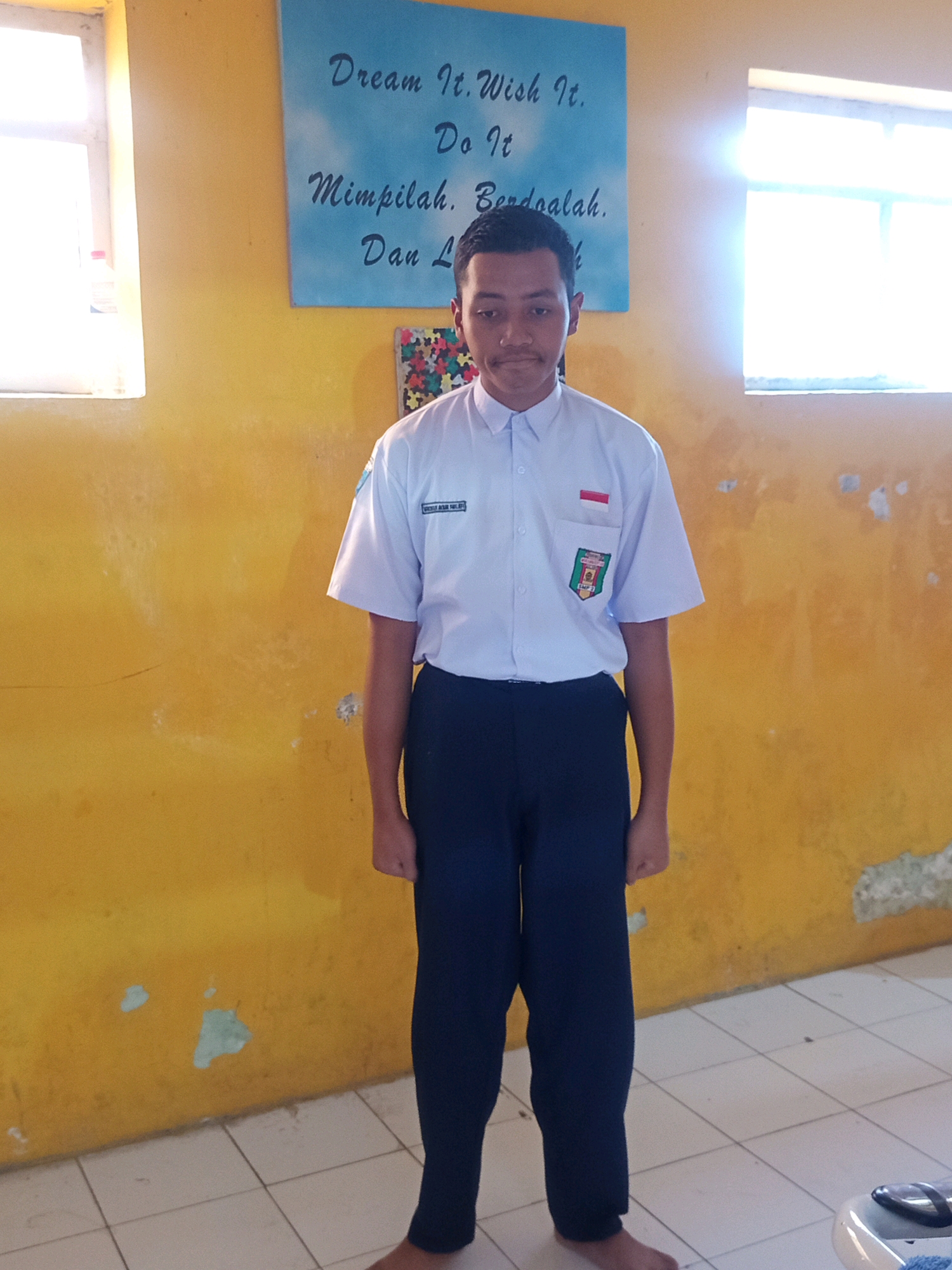 A young Indonesian boy stands in front of a yellow wall with a motivational poster in the background. He is wearing a white, short-sleeved school uniform shirt with a crest on the left breast and a small Indonesian flag on the right breast. He is also wearing dark blue pants. The poster behind him has text in English and Indonesian, reading "Dream It. Wish It. Do It." and "Mimpilah. Berdoalah. Dan Lakukan." to the right of that. There are two windows in the background, one on the left and one on the right, both letting in bright sunlight. The floor is tiled with white square tiles. The wall appears to be painted yellow and shows some signs of wear and tear, with patches of peeling paint. The boy's face is looking directly at the camera with a neutral expression. The overall scene appears to be indoors, possibly a classroom or hallway, in the city of Malang, Indonesia.