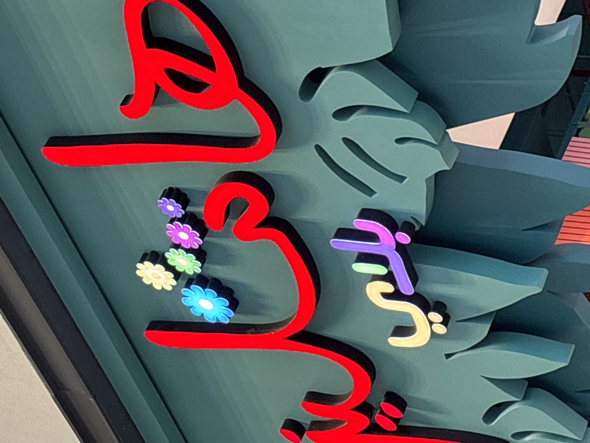 The image displays a close-up, eye-level view of a neon sign in Arabic script, featuring stylized flowers and leaf-like decorative elements. The sign is mounted on a teal-colored wall. The Arabic lettering is in a vibrant red, outlined with black. Below the main red lettering, there is a smaller sign with different colored letters. The leftmost characters of the smaller sign are in yellow and purple. The overall aesthetic suggests a playful and possibly commercial establishment. The image is cropped to focus on the sign itself, with no discernible background elements that would indicate a specific location within Dubai, United Arab Emirates.