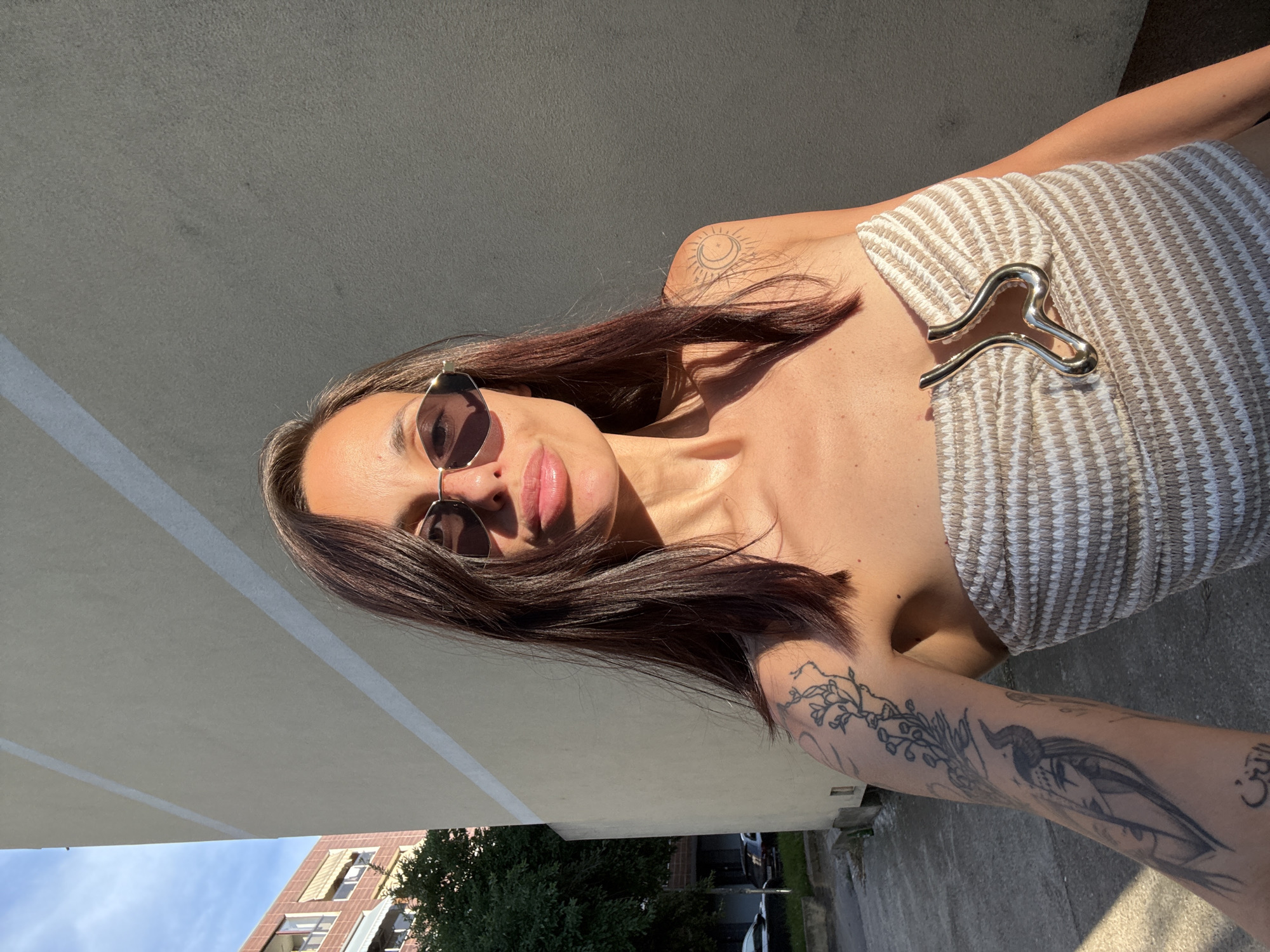 A young woman with shoulder-length dark brown hair, wearing sunglasses and a strapless beige and white striped top with a silver metallic detail, poses for a selfie outdoors. She has visible tattoos on her left arm and shoulder, including a floral design and a sun and moon symbol. The background features a textured beige wall with horizontal white stripes, a building with balconies, trees, and a street with parked cars. The lighting suggests it is late afternoon, with golden sunlight casting shadows. The overall scene depicts a casual outdoor moment in an urban environment. The location is identified as Pleven, Bulgaria.