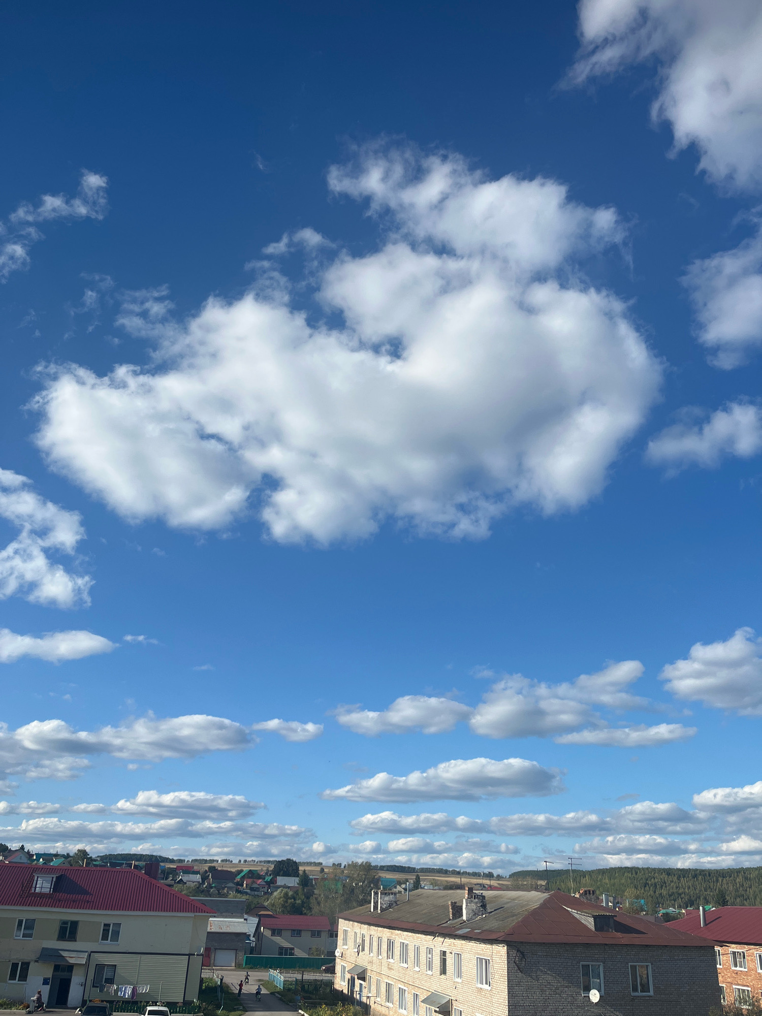 The image is a daytime shot of a residential area in Bizhbulyak, Russia, under a bright blue sky filled with fluffy white clouds. In the foreground, there are several buildings with red and gray roofs. One building on the left appears to be a multi-story apartment building with light-colored walls. Clothes are hanging on a line outside of it. To the right, a brick building with a gray roof and multiple windows is visible. Further into the distance, more houses and buildings with colorful roofs are scattered throughout the landscape, with a line of trees and hills in the background. The scene appears calm and ordinary, with no obvious signs of distress or unusual activity.