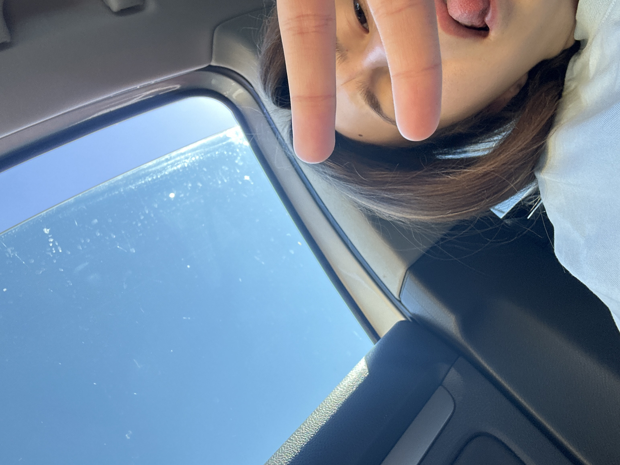 A young woman with long, dark hair sticks her tongue out at the camera while making a peace sign with her fingers over her right eye. She is wearing a light-colored top and is inside a vehicle, as indicated by the car's interior visible in the foreground. The sun is shining brightly through the car's sunroof, illuminating the sky and the woman's face. The image appears to have been taken during the daytime in Shumen, Bulgaria.