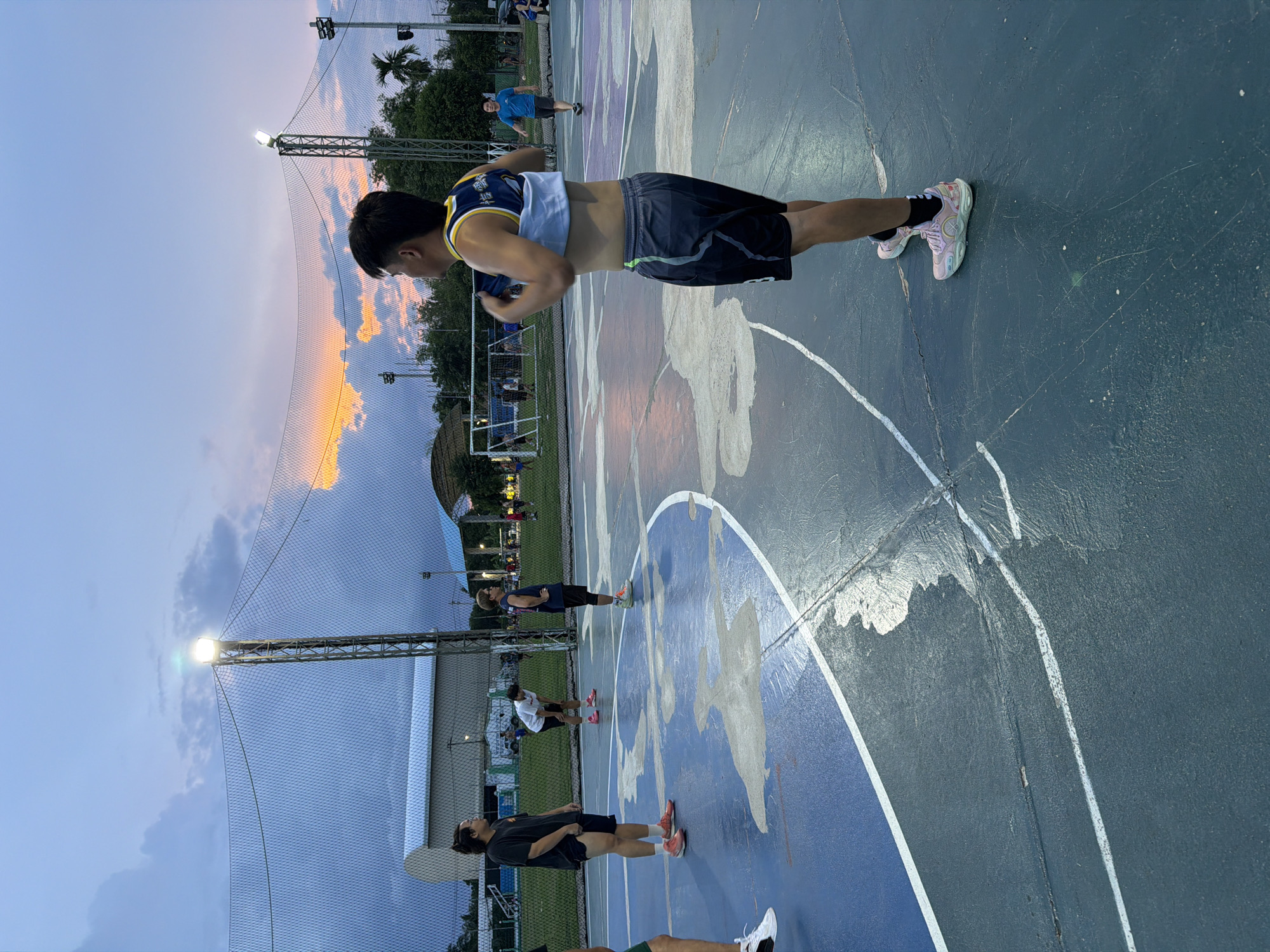 The image shows a group of people exercising outdoors on a basketball court in Chang Phueak, Thailand. The court has a blue and grey surface with white lines and painted designs. Several individuals are seen performing various exercises. A man in the foreground is doing push-ups. Others are stretching or engaging in similar activities. The background includes trees, a building, and a sunset sky. The court is surrounded by a net fence and illuminated by bright lights.