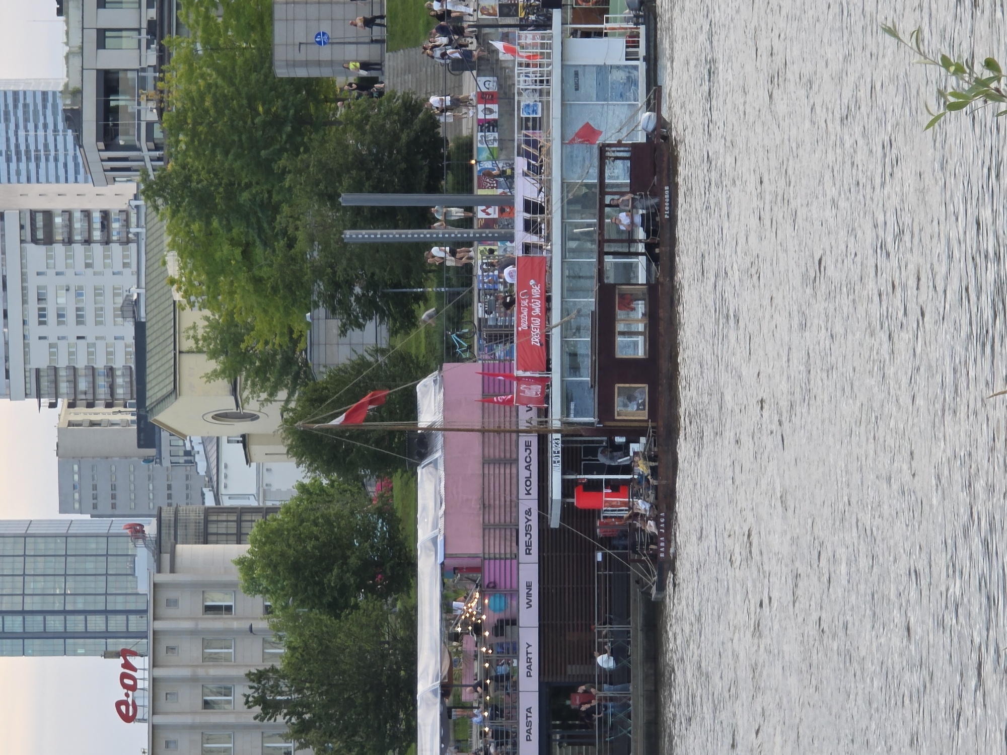 The image shows a view of the Vistula River in Warsaw, Poland, with several houseboats and buildings visible along the embankment. In the foreground, the water has ripples and a slight sheen, suggesting it is daytime.
The most prominent subject is a multi-level houseboat or floating venue, painted in shades of pink and white, with a large, transparent canopy covering the upper deck. It is adorned with numerous string lights and several red flags. A Polish flag is also visible flying from a mast on the boat. A banner across the boat reads "Zresetuj się zresetuj swój vibe". Another section of the boat displays text including "PASTA PARTY WINE REJSY & KOLACJE". A smaller, older-looking houseboat is moored alongside, with the name "BABA JAGA" visible on its stern. Several people are visible on the decks of both boats, some appearing to be customers and others staff.
Behind the houseboats, a row of trees forms a green backdrop. Beyond the trees, a variety of modern and older buildings are visible. Notable structures include a tall, glass-fronted skyscraper with the red "e-on" logo at its base, and several other multi-story buildings of varying architectural styles, some with visible windows and balconies. The sky above is overcast, with a pale, diffused light. Stairs and a paved area lead up from the embankment, with several people standing and walking on them.