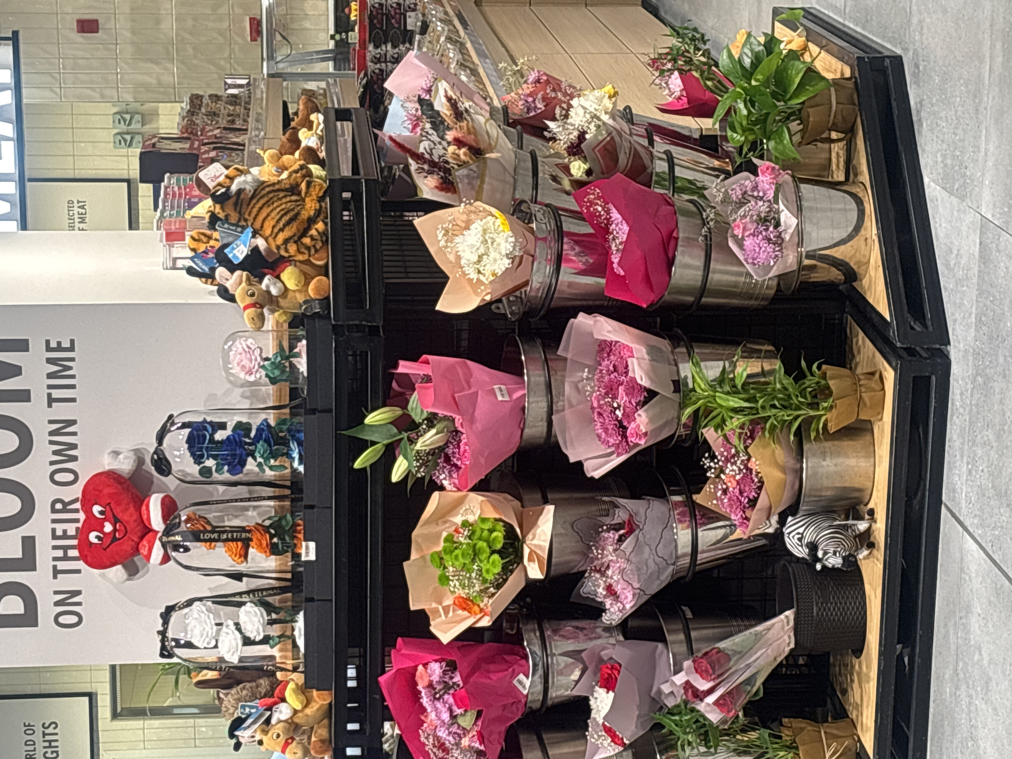 The image depicts a retail display of flowers and plush toys in what appears to be a supermarket or a flower shop. The display features several arrangements of fresh flowers in buckets, wrapped in colorful paper. Among the flowers, there are also plush toys, including a tiger, a camel, and teddy bears, placed on shelves above the flowers. In the foreground, a small zebra figurine is positioned near a bucket of flowers. The background shows shelves stocked with various products, suggesting a commercial setting. The signage visible in the background indicates the location as Dubai, United Arab Emirates, with text related to "MEAT" and "SELECTED MEAT." The overall scene is well-lit and organized, characteristic of a retail environment. There are no people visible in the image.