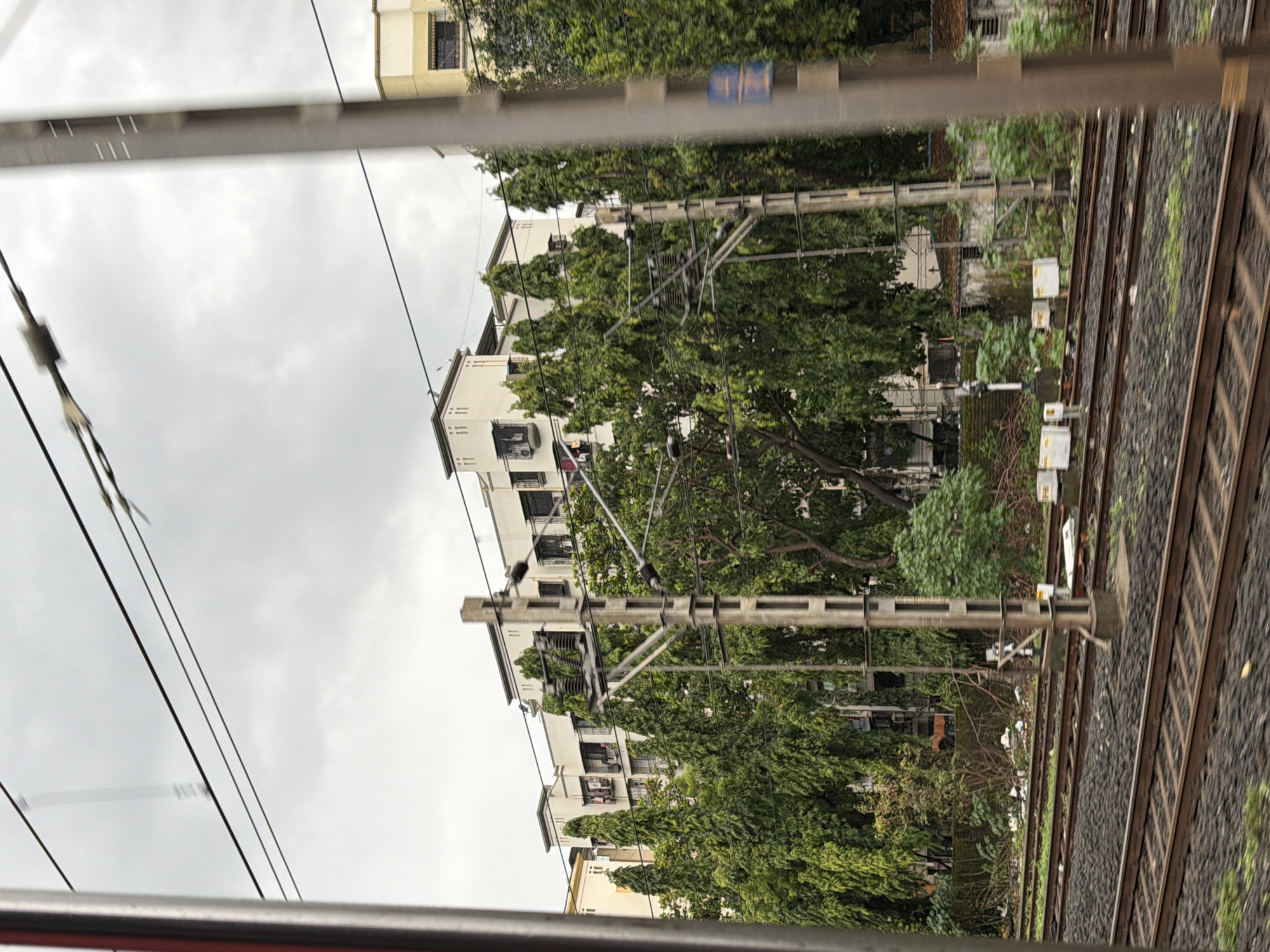 The image captures a view from a moving train, likely on a cloudy day, in Mira-Bhayander, India. The foreground shows multiple sets of railway tracks, with dark ballast and some green vegetation growing between them. A tall, grey electrical pylon with various insulators and wires is prominently featured on the right side of the frame. In the middle ground, a dense line of tall, conical green trees obscures much of the view. Behind the trees, several multi-story apartment buildings with white facades are visible. These buildings have multiple windows, balconies, and some have air conditioning units. The sky above is overcast with a light grey color. The overall impression is an urban or suburban environment with railway infrastructure, residential buildings, and abundant greenery.