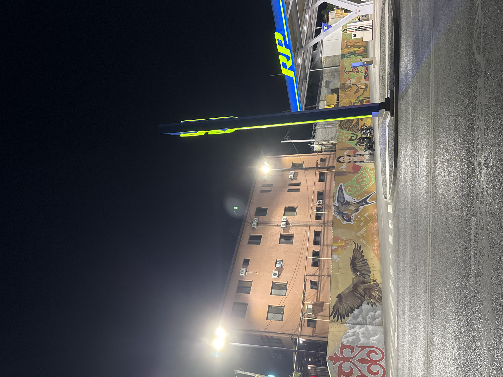 The image depicts a street scene at night, possibly in Shymkent, Kazakhstan, illuminated by artificial lights. A prominent blue and yellow structure, likely a gas station canopy, dominates the upper portion of the image, featuring illuminated signage with the letters "RP" in yellow. Below this structure, a multi-story building with a peach-colored facade is visible, adorned with large-scale murals. These murals feature an eagle in flight against a cloudy background, a swallow, and decorative patterns. The building has numerous windows and air conditioning units. A wet asphalt road runs alongside the building, reflecting the lights. The overall atmosphere suggests a quiet urban environment at night, with a focus on the architecture and street art. The presence of streetlights and the illuminated gas station canopy indicate that it is nighttime. The wet road suggests recent rain or dew. No people are clearly visible in the frame.