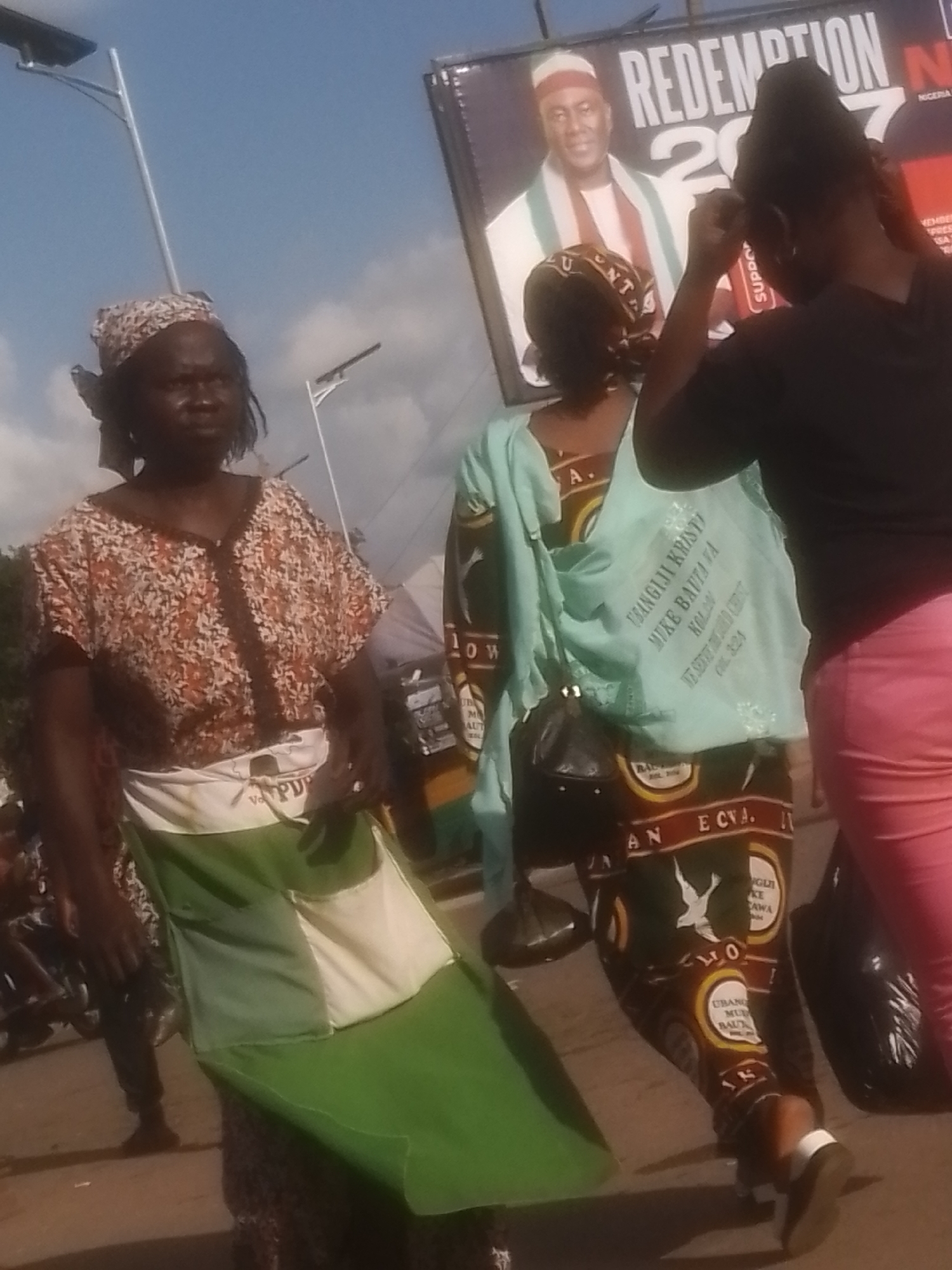 In Gwong, Nigeria, three women stand on a paved surface. The woman on the left wears a patterned headscarf and a patterned brown and white top, with a white and green Nigerian flag draped over her front. She has dark skin and is looking to her right. In the center, a woman in a light blue headscarf and matching light blue garment stands with her back to the camera. She carries a dark purse. Her garment has writing on it. On the right, a woman with dark skin wears a black top and pink pants, facing away from the camera. She is adjusting her hair with her right hand. Behind them, a large billboard features a portrait of a man wearing a white and green outfit, with the word "REDEMPTION" and "2017" visible. The sky is blue with some clouds. Streetlights are visible in the background.