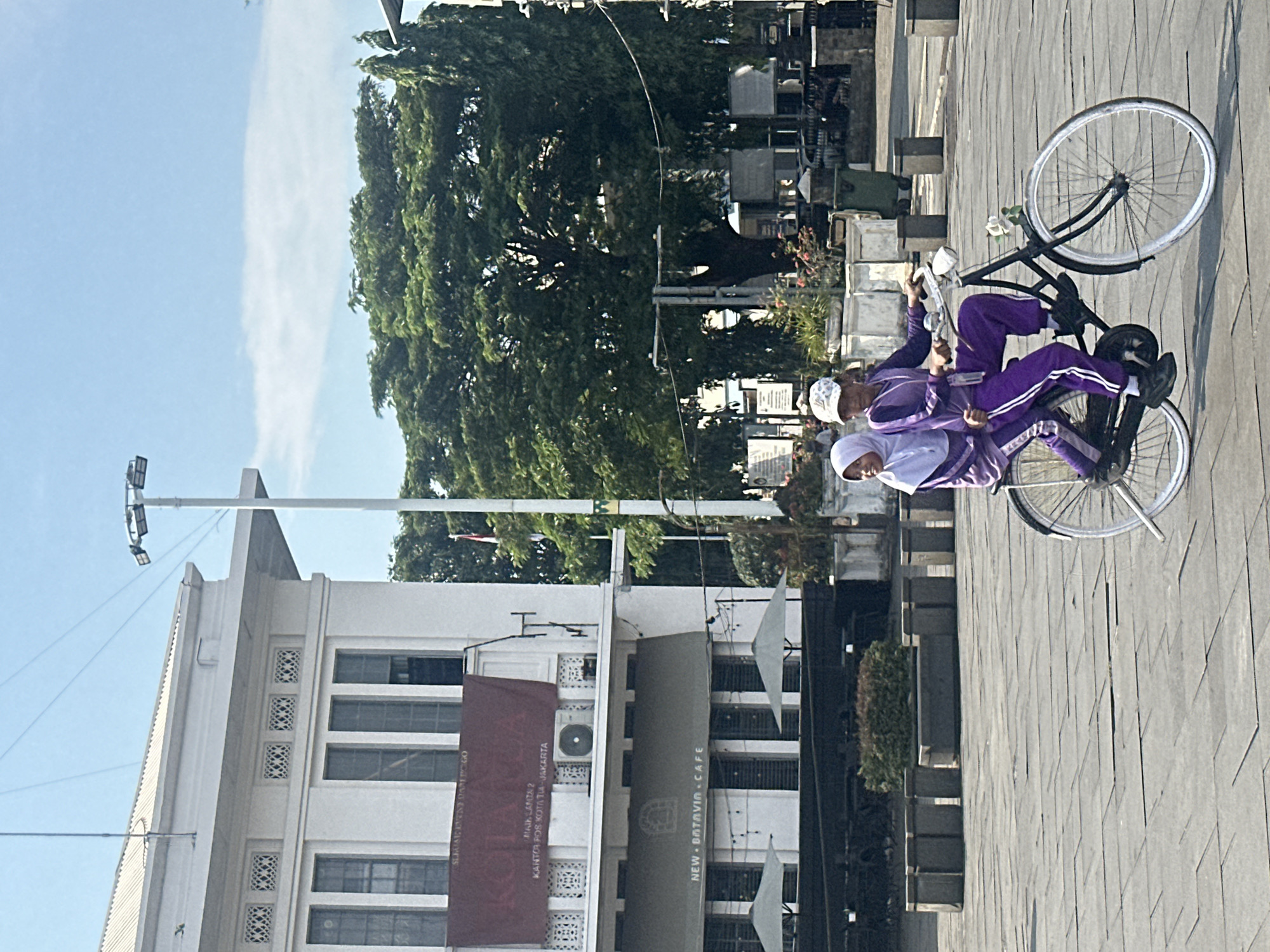 Two people, presumably children, are riding a bicycle on a cobblestone-paved street in West Jakarta, Indonesia. The person in the front is wearing a white cap and the person in the back is wearing a patterned cap. Both are wearing purple tracksuits. Behind them is a building with a sign for "NEW BATAVIN.CAFE". There is also a red banner indicating "MAIL LAMTA 2, KANTOR POS KOTA TLA-JAKARTA". A tall tree is visible behind the building, and the sky is light blue with a patch of white clouds. A light pole stands to the left of the building.