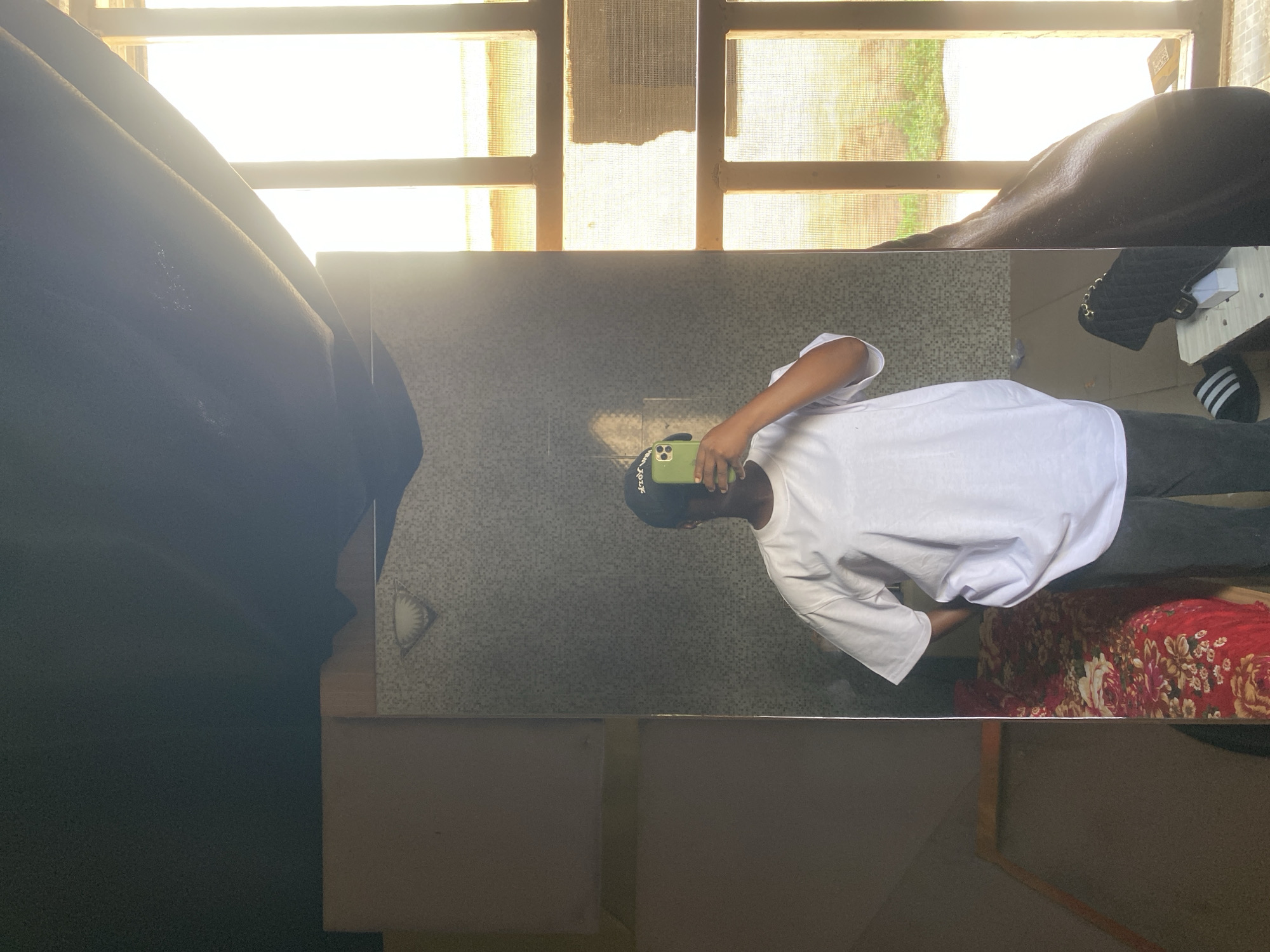 A person is taking a selfie in front of a mirror. The person is wearing a white shirt, dark pants, and a dark hat. A smartphone is in the person's hand. The mirror is reflecting the person, as well as the background. The background includes a window with blinds, a dark curtain, a patterned blanket, and a bag. The location is Unknown, Unknown.