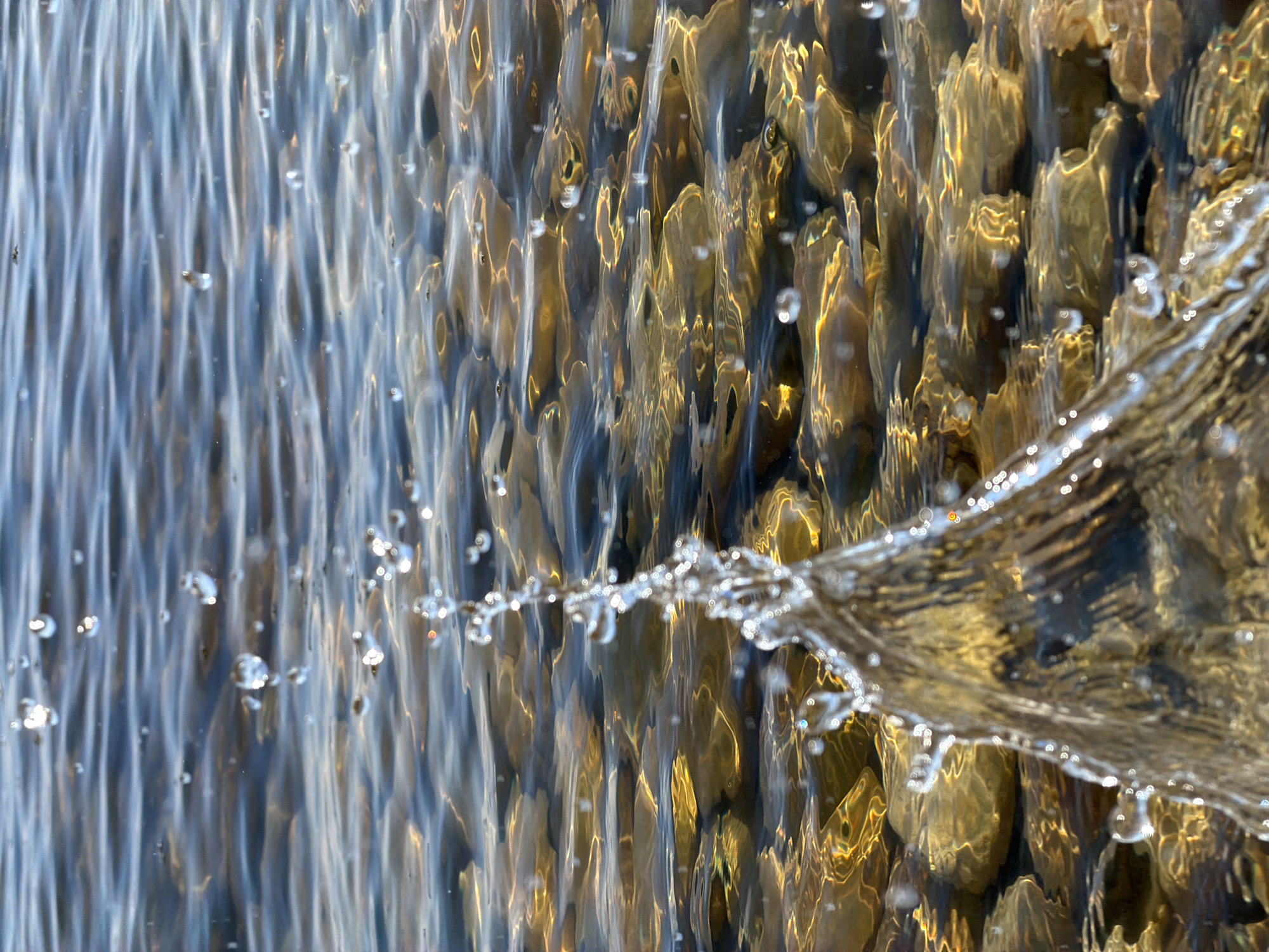 A close-up captures a cascading waterfall. The water is clear, with droplets suspended in the air. The background reveals amber and brown stones. The location is Kamyshin, Russia.