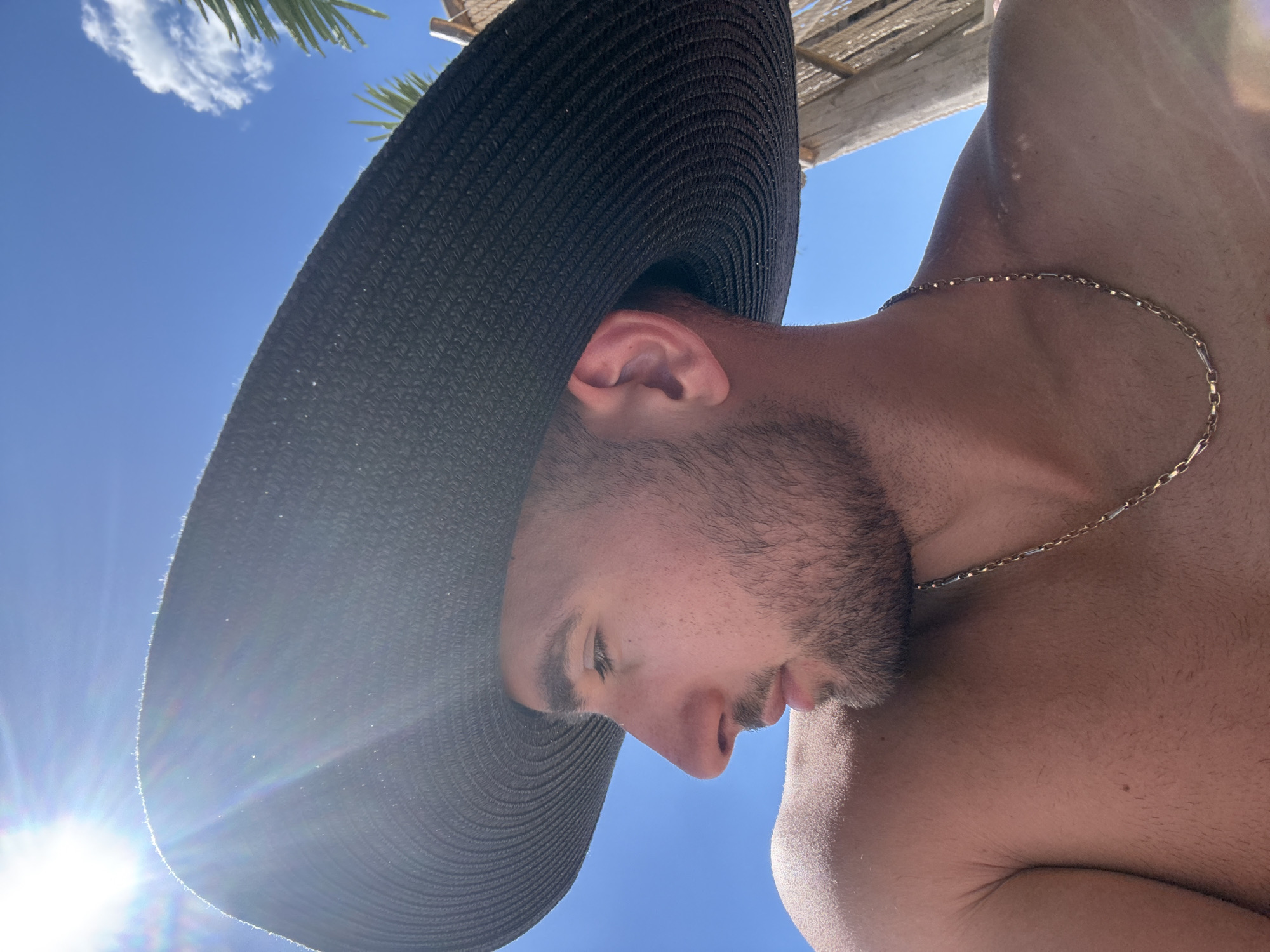 The image shows a close-up, profile view of a young man wearing a large, dark-brimmed hat. He is shirtless, and a gold chain is visible around his neck. The man is looking to his left, away from the camera. The background is a clear blue sky with the sun shining brightly, creating lens flare. Some palm fronds and a wooden structure are visible on the right side of the image. The man appears to be outdoors, possibly at a beach or resort.
There is no information in the image that allows for the specific determination of the location being the city of Nesebar, Bulgaria.