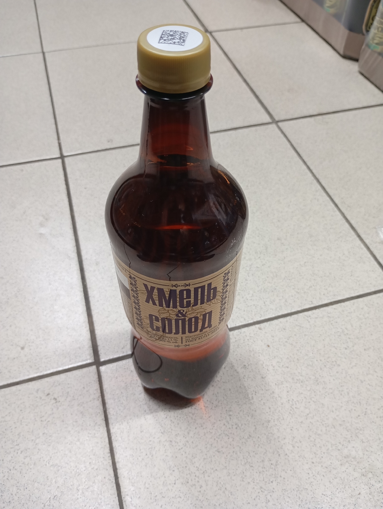 A brown plastic bottle with a gold cap sits on a light gray tiled floor. The bottle has a label with Cyrillic text and a QR code on the cap. The background is blurred, but appears to be a store aisle with shelves of products. The lighting suggests an indoor commercial setting. No people or specific Moscow landmarks are visible.