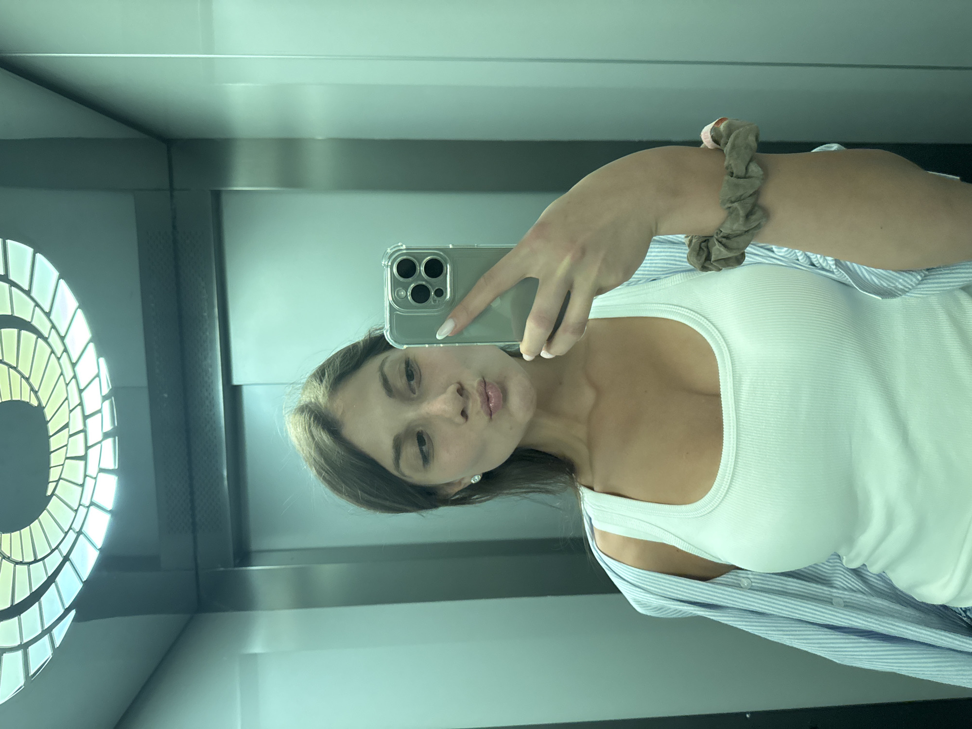 The image shows a young woman taking a mirror selfie in what appears to be an elevator. She is wearing a white tank top, a light blue and white striped unbuttoned shirt, and a scrunchie on her left arm. The elevator has metallic walls and a decorative ceiling light. The woman is puckering her lips for the selfie. The image is taken from a medium shot, focusing on the woman and her immediate surroundings within the elevator.