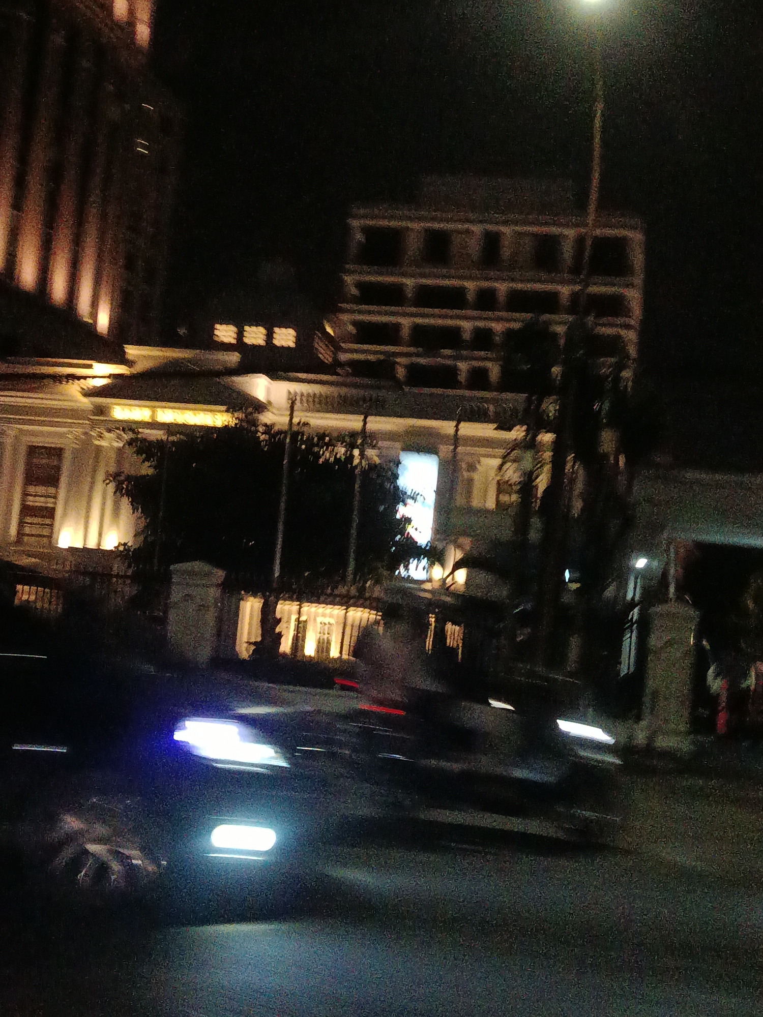 The image shows a street scene at night in the city of Medan, Indonesia. A multi-story building with lit windows is visible on the right side of the frame, with a tall pole extending upwards, possibly a lamppost. To the left, another building with a classical architectural style is illuminated by warm light from its windows and facade. In the foreground, the headlights of a vehicle are blurred due to motion, indicating it is either moving or the camera is panning. The street is dark, and other vehicles or figures are indistinct due to low light and motion blur. The overall impression is a nocturnal urban environment with illuminated architecture and moving traffic.
