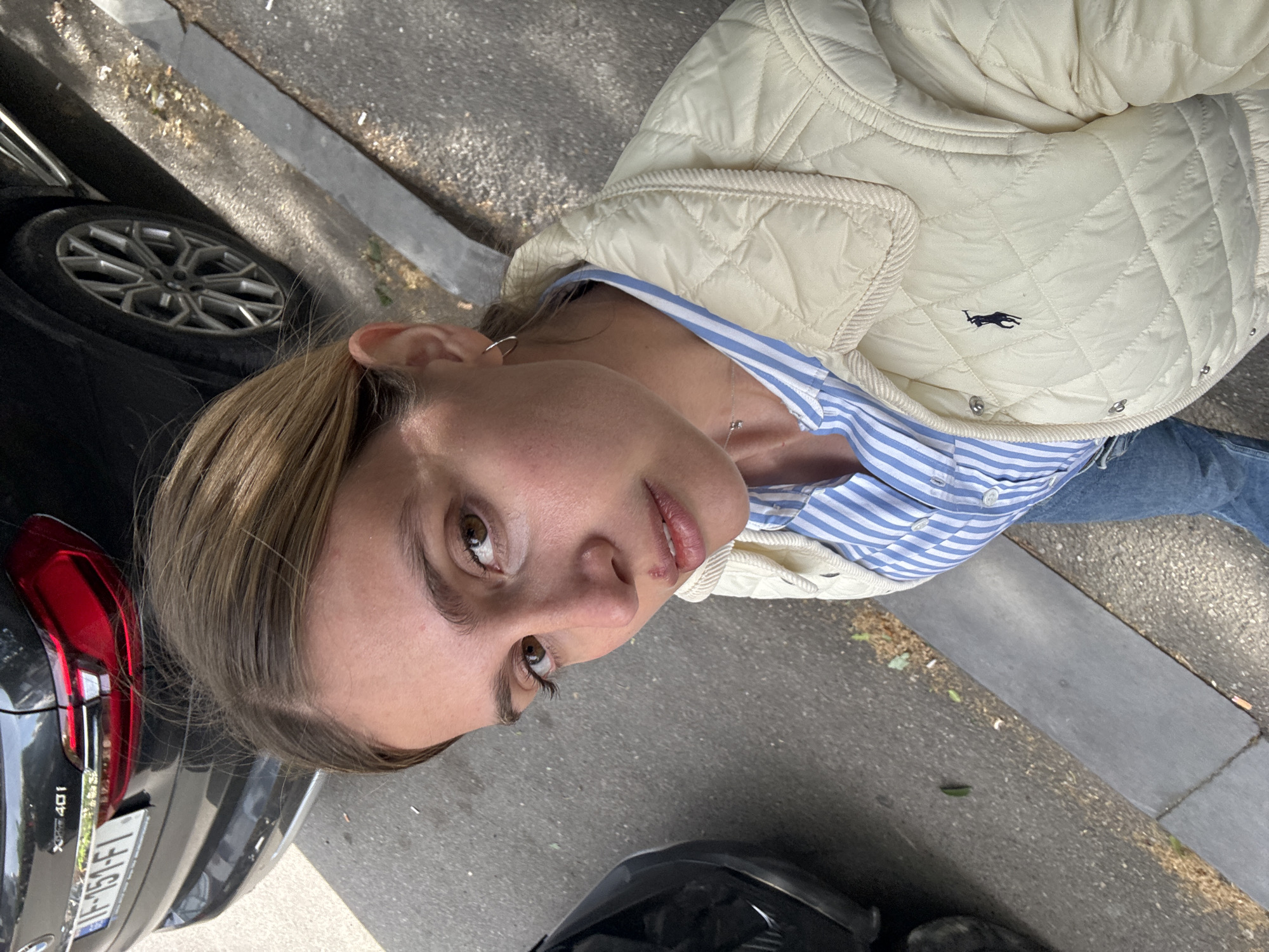 A young woman is the central subject of this close-up, angled photograph, appearing to be on a city street during the daytime. She looks upwards and slightly towards the camera with a neutral to slightly contemplative expression. Her light brown hair is parted on the side, framing her face with brown eyes. She is wearing a light-colored, possibly cream or off-white, quilted jacket that features a small, dark embroidered logo of a polo player on a horse, characteristic of the Ralph Lauren brand. Underneath the jacket, she has a blue and white striped collared shirt, and she is also wearing blue jeans, partially visible. A delicate silver necklace with a tiny pendant hangs around her neck, and a small silver hoop earring is visible in her left ear.
In the background, a dark-colored car, likely an SUV, is parked on the left side of the frame. Its red and black taillight, a portion of its wheel with a distinctive rim design, and the text "XDRIVE 40I" indicating a BMW model are visible. The car's license plate reads "IF-151-FI". The setting shows an asphalt road, a grey curb, and a textured paved area, suggesting an urban environment. The bright illumination indicates it is daytime. Another dark object, possibly part of a vehicle, is in the lower right foreground. The scene captures a casual street moment in Tbilisi, Georgia.