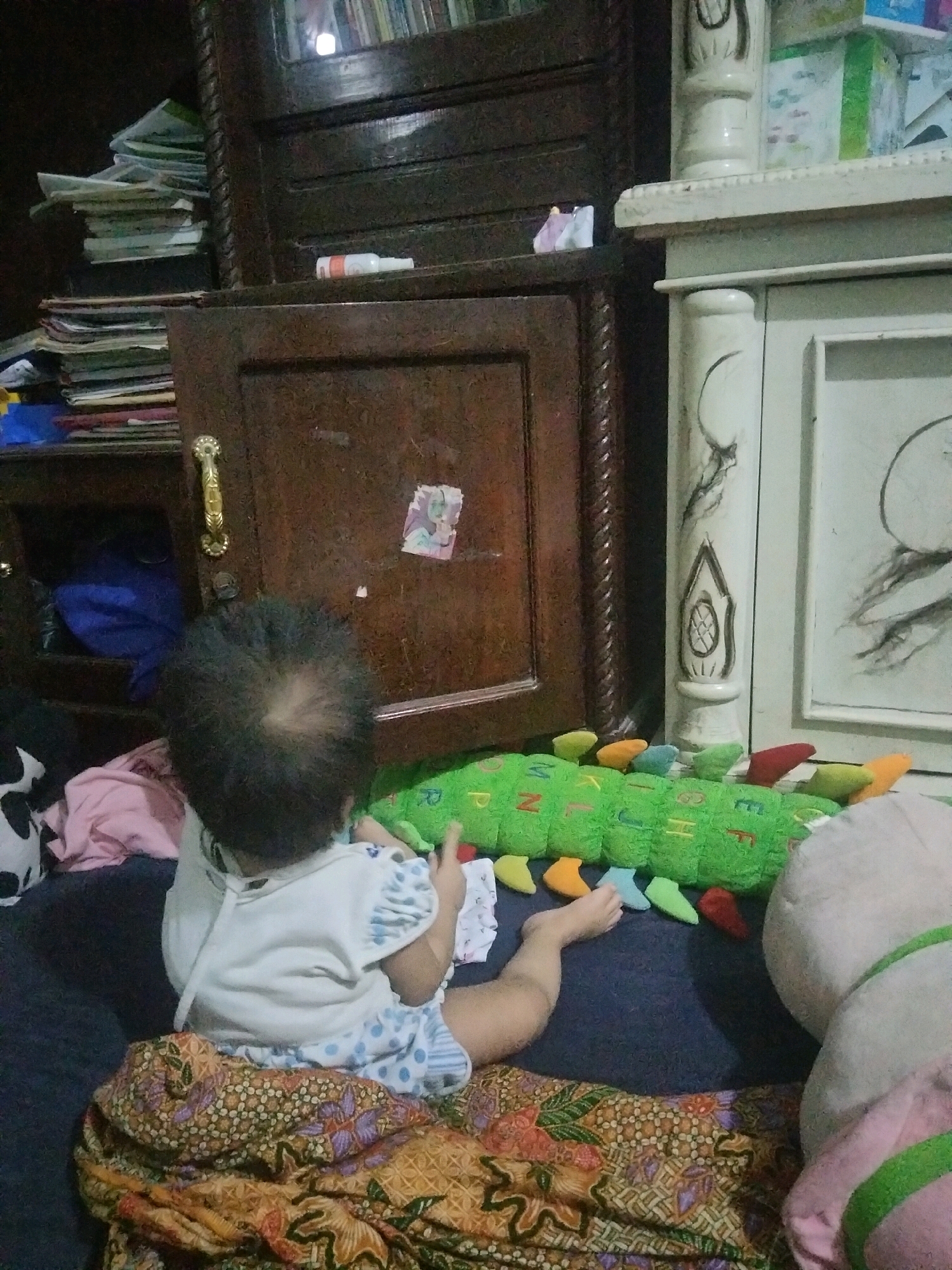 In a room in Suka Sari, Indonesia, a toddler sits on the floor facing away from the camera. The child wears a white shirt with blue polka dots and a white bib. A large, green, caterpillar-shaped stuffed toy with letters on it lies in front of the child. A patterned blanket is spread out on the floor. In the background, a wooden cabinet with a gold handle is visible, along with a white decorative pillar. The environment appears to be a living space within a home.