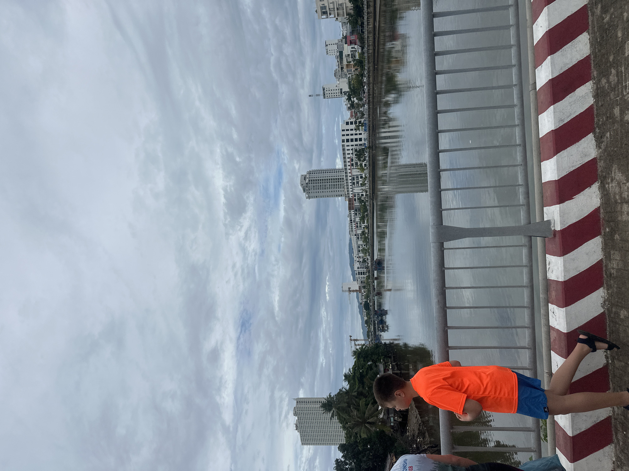 A medium, eye-level, outdoor shot shows a boy looking over a metal bridge railing along a waterway. The boy, wearing an orange shirt and blue shorts, is looking out over the water. Part of another person wearing a gray shirt can be seen partially in the lower foreground. A red and white stripe-painted curb runs along the bridge rail on the right side of the photo. In the background are multiple buildings, some are reflected in the water. The sky is overcast with many clouds. The location is Nha Trang Ward, Vietnam.