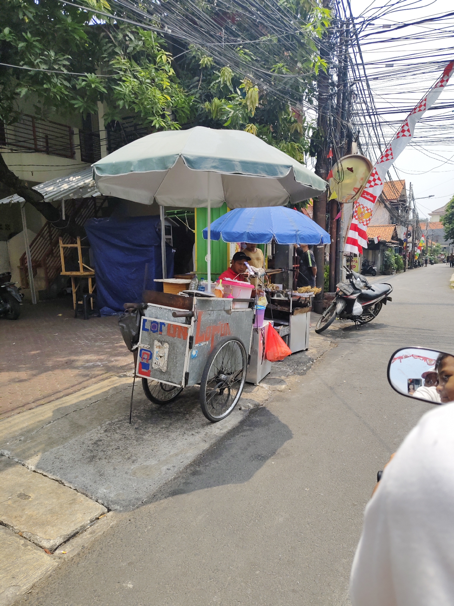 The image depicts a street scene in South Jakarta, Indonesia. In the foreground, a food cart with the word "LORONG" visible on its side is parked on the side of a paved road. The cart has two wheels and appears to be made of metal. It is covered by two umbrellas, one large and white, and a smaller blue one. A person wearing a red shirt is tending to the cart, and another person is visible in the background.
To the left of the cart, there is a building with a balcony and some trees. Various electrical wires are strung across the scene, typical of urban environments in Indonesia. A red and white Indonesian flag is also visible on a pole. Several motorcycles are parked along the street, and a few people are walking in the distance. The scene is illuminated by sunlight, casting shadows on the road. The image appears to have been taken from the perspective of someone riding a motorcycle.