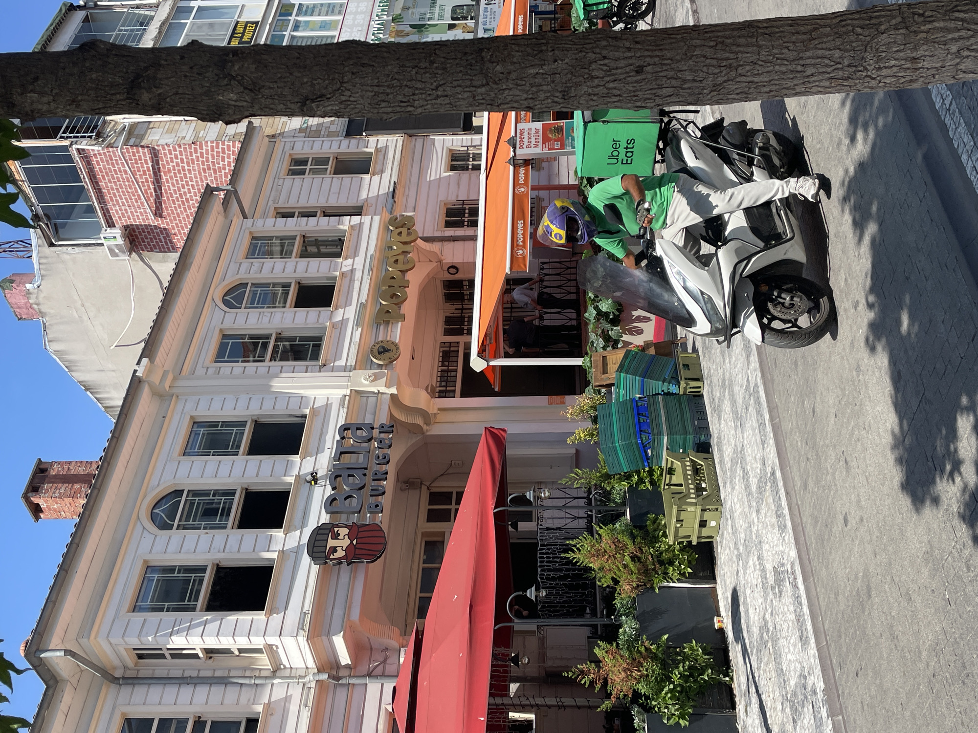 The image depicts a street scene in the city of Kadıköy, Turkey, with a focus on the facades of several businesses and a delivery person on a scooter. The foreground is dominated by a white scooter ridden by a person wearing a green shirt, light-colored pants, white sneakers, and a purple and yellow helmet. The scooter has a large green Uber Eats delivery box mounted on the rear. The rider's left hand is on the handlebars, and their right hand is holding the front brake lever.
Behind the scooter, two restaurants are visible. The one on the left has a sign that reads "Balta Burger" with a cartoonish character featuring a beard and a hat. This establishment has an outdoor seating area covered by a large red umbrella, with potted plants and stacked green plastic crates in front of it. The restaurant to its right has an orange awning and a sign that says "Popeyes." People can be seen standing under the awning of Popeyes.
The buildings have a traditional architectural style with white facades, decorative window frames, and exposed brickwork on higher levels. In the background, there are other buildings, including a multi-story structure with balconies and a large tree trunk partially obscures the right side of the image. The street is paved with interlocking stones and asphalt. The sky is clear and blue, indicating a sunny day.