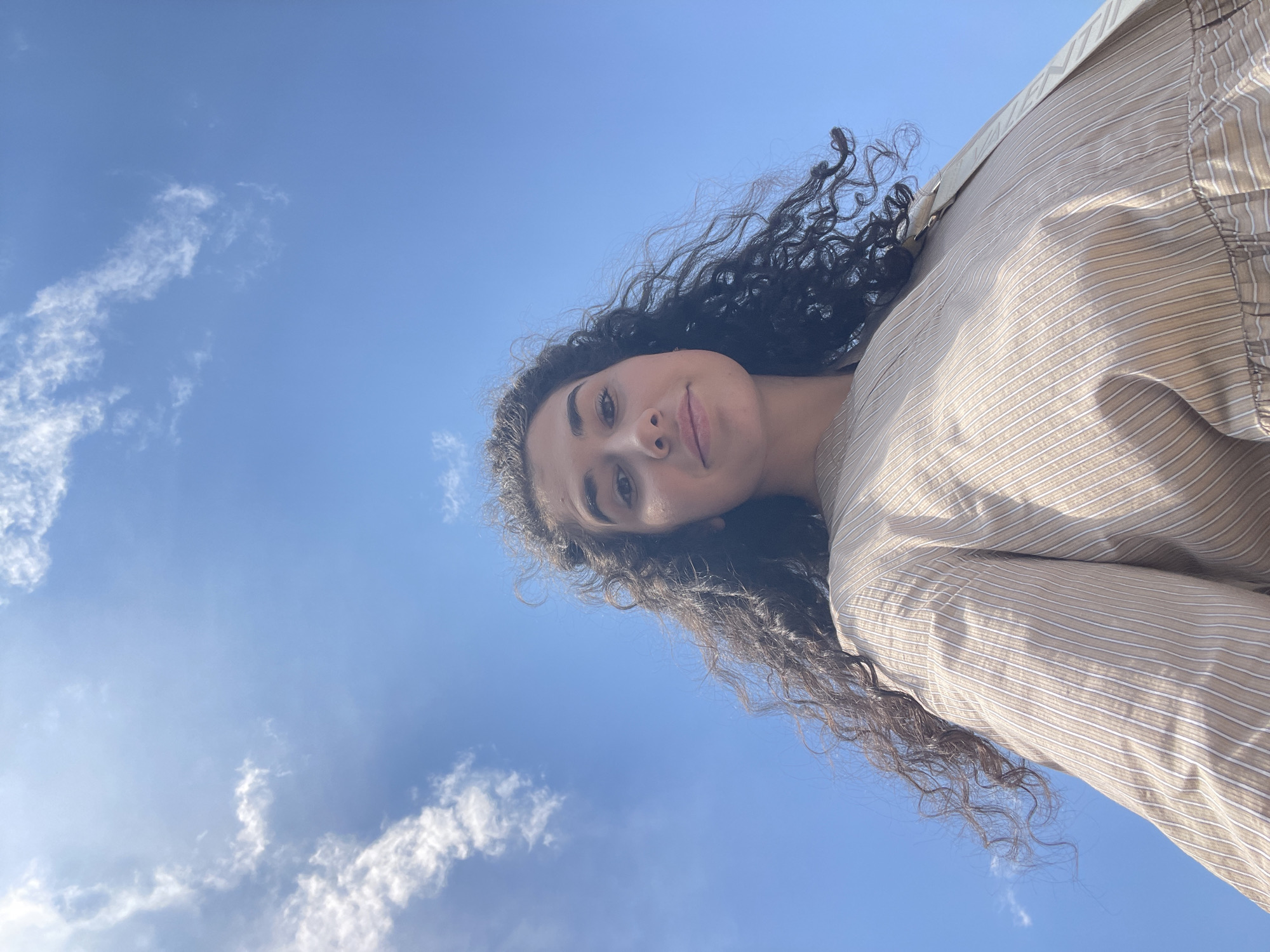 A low-angle shot captures a young woman from the chest up, her curly dark hair flowing around her face. She is wearing a light brown and white striped button-up shirt, which is slightly off-center in the frame. The background is a clear blue sky with a few wispy white clouds, suggesting an outdoor setting. The perspective of the photograph, looking up at the woman against the sky, creates a sense of openness and perhaps a bit of drama. The lighting suggests it is daytime. There is no identifiable location in the city of Sofia, Bulgaria.