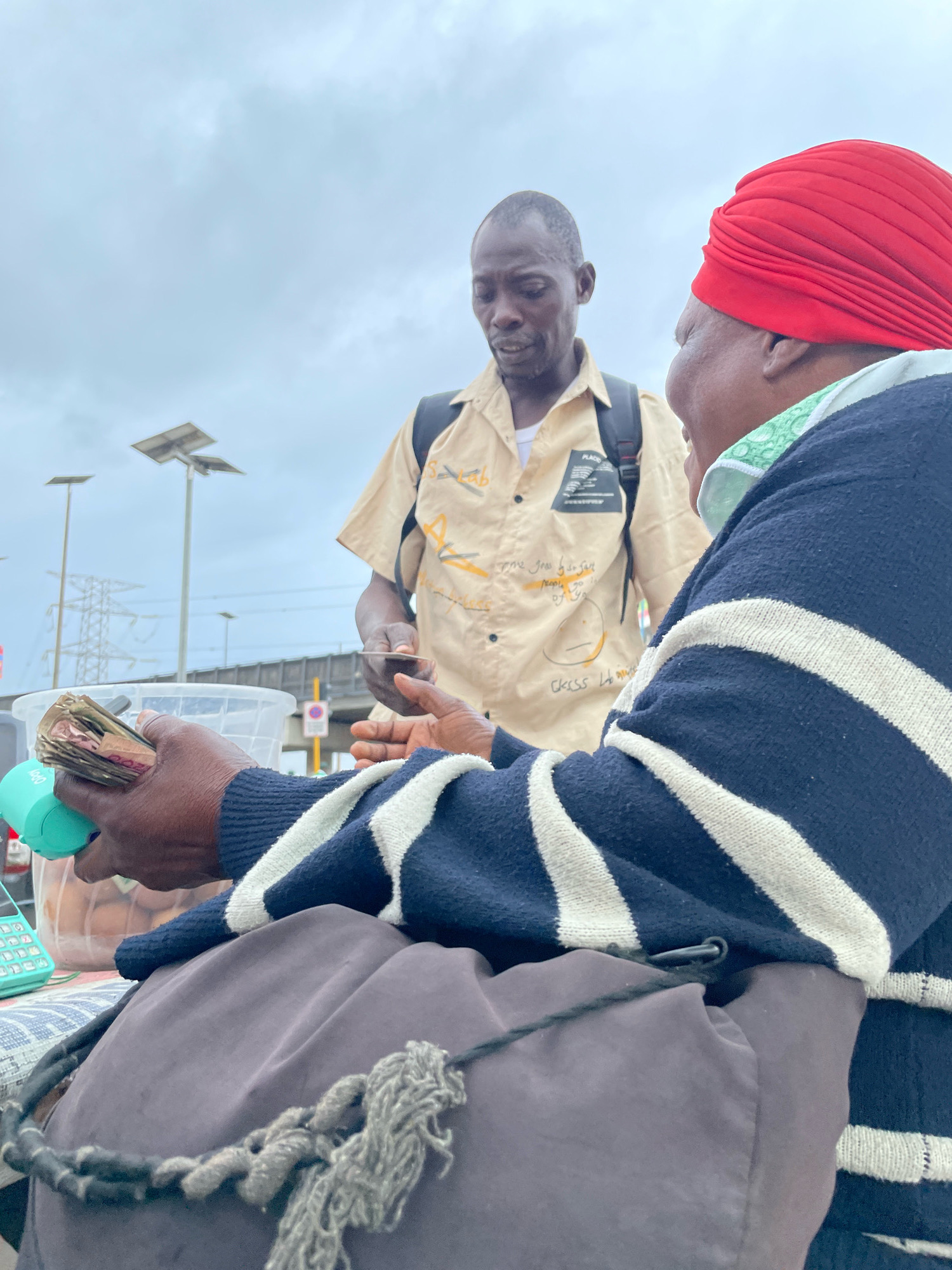 In Ajah, Nigeria, on a cloudy day, a transaction is occurring between two individuals. In the foreground, a person wearing a navy blue and white striped sweater and a grey skirt holds a wad of cash and a teal-colored object. Next to them, a calculator and a clear plastic container filled with eggs are visible. The second individual, standing behind, is wearing a tan shirt with markings and a black backpack, and appears to be handing a small object, possibly another piece of currency or a card, to the person in the foreground. The background shows power lines, streetlights, and a bridge, suggesting an urban environment. The people's facial expressions are obscured or not fully visible, focusing the attention on the exchange.