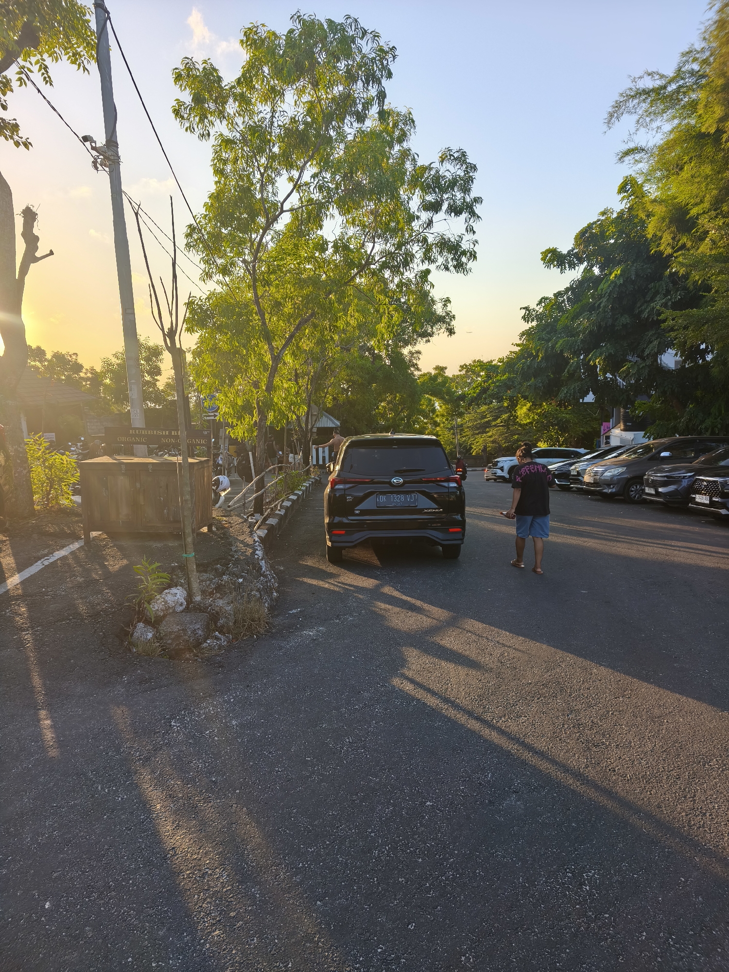 An outdoor scene in Kuta Selatan, Indonesia, captures a parking area bathed in the warm, low light of the setting sun. The sky transitions from a clear, pale blue at the top to a soft yellow near the horizon, with a single wispy cloud visible. Tall trees with dense green foliage line the right side of the frame, their shadows stretching long and distinct across the asphalt. On the left, thinner trees and a utility pole are silhouetted against the bright sky. A black SUV is parked in the foreground, its license plate displaying "DK 1328 VJ." A person wearing a black t-shirt with a graphic design and blue shorts walks away from the camera on the asphalt. In the background, several other cars are parked, and a wooden structure with signage, possibly a recycling bin area, is visible. The ground is a dark asphalt surface with scattered rocks and vegetation along the edge.