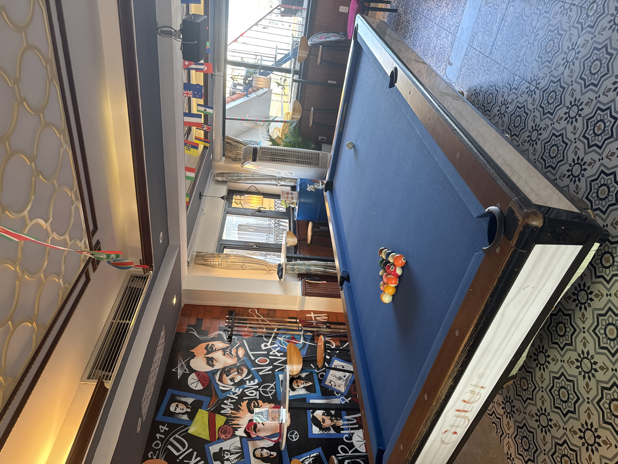 This image depicts a pool table in a room in Hội An Ward, Vietnam. The table is set up with a racked set of pool balls and a cue ball already on the blue playing surface. A set of pool cues leans against the wall beside the table. The floor has a geometric tile pattern. The walls are decorated with murals and flags. Outside of a window in the background, another level of seating can be seen with bar stools.
