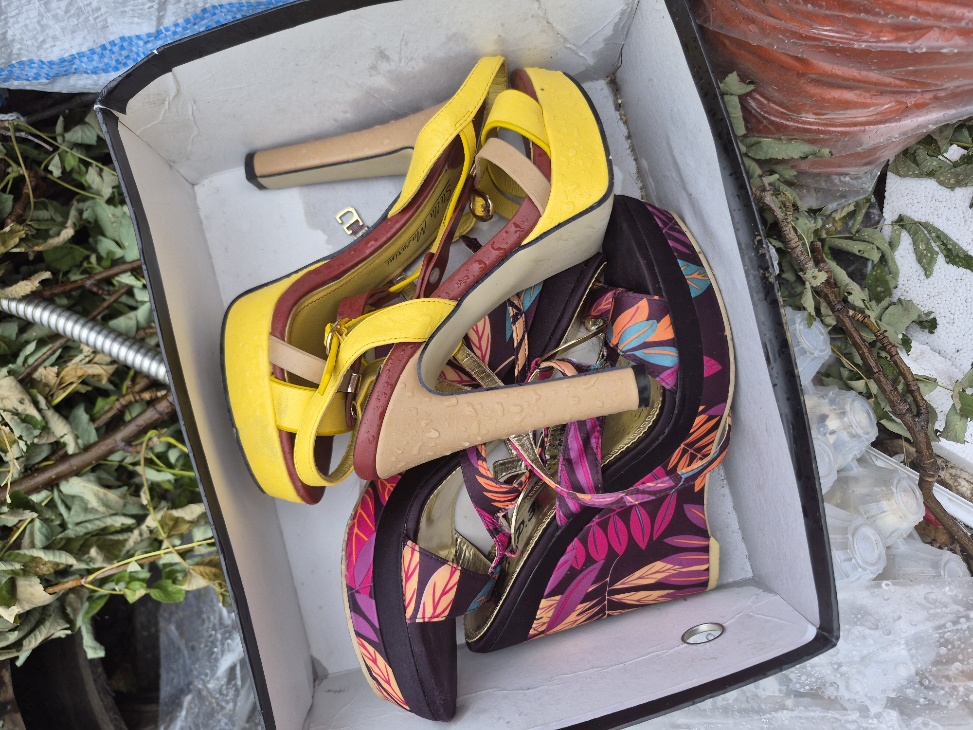 A cardboard box filled with assorted women's high-heeled shoes is shown. The shoes are a mix of styles and colors, including a bright yellow pair with red and tan accents, and another pair with a dark purple base and a vibrant floral pattern in shades of pink, orange, and blue. The shoes appear to be damp, suggesting they may have been exposed to rain or water. The box is open and situated outdoors, surrounded by fallen leaves, branches, and some plastic debris, hinting at a discarded or outdoor setting. In the background, out of focus, is a roll of orange material wrapped in plastic. The overall impression is one of discarded items in an outdoor environment, possibly after a period of wet weather. No people are visible in the image. The visible text on the yellow shoe reads "Stella Mazarini". The setting appears to be in Nyagan, Russia.