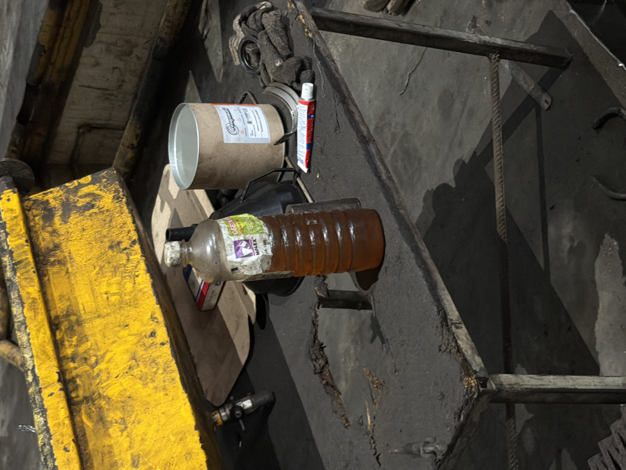 A bottle of amber liquid with a white label is positioned on a dirty metal table. Next to the bottle is a brown cardboard cylinder with a label. There is also a red and white tube lying on the table. In the background, a large yellow piece of machinery is visible. The scene appears to be an industrial or workshop environment, possibly located in Oktyabrsky, Russia.