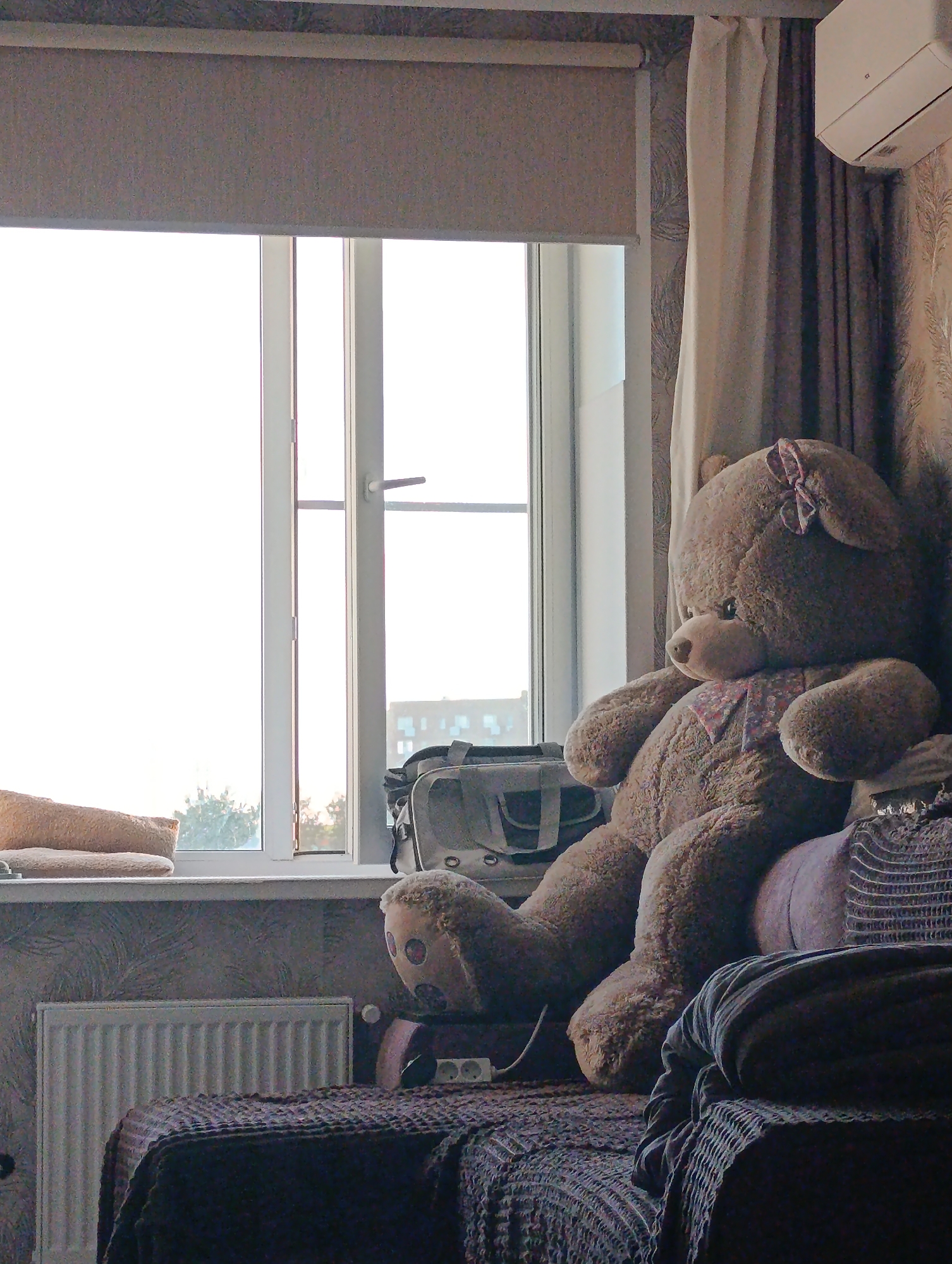 A large, fuzzy, brown teddy bear with a pink bow on its head sits on a bed next to a window. The teddy bear is wearing a small, patterned bandana around its neck. The window is large and appears to be a sliding glass door, looking out onto trees and buildings. A roller blind is partially drawn over the top of the window. To the left of the teddy bear, on the windowsill, is a tan-colored pillow. Below the window is a white radiator. The bed is covered with dark-colored blankets and pillows, and a part of another blanket is draped over the edge of the bed in the foreground. A grey bag with straps is placed behind the teddy bear. The room is decorated with wallpaper. The scene is well-lit by natural light coming from the window.