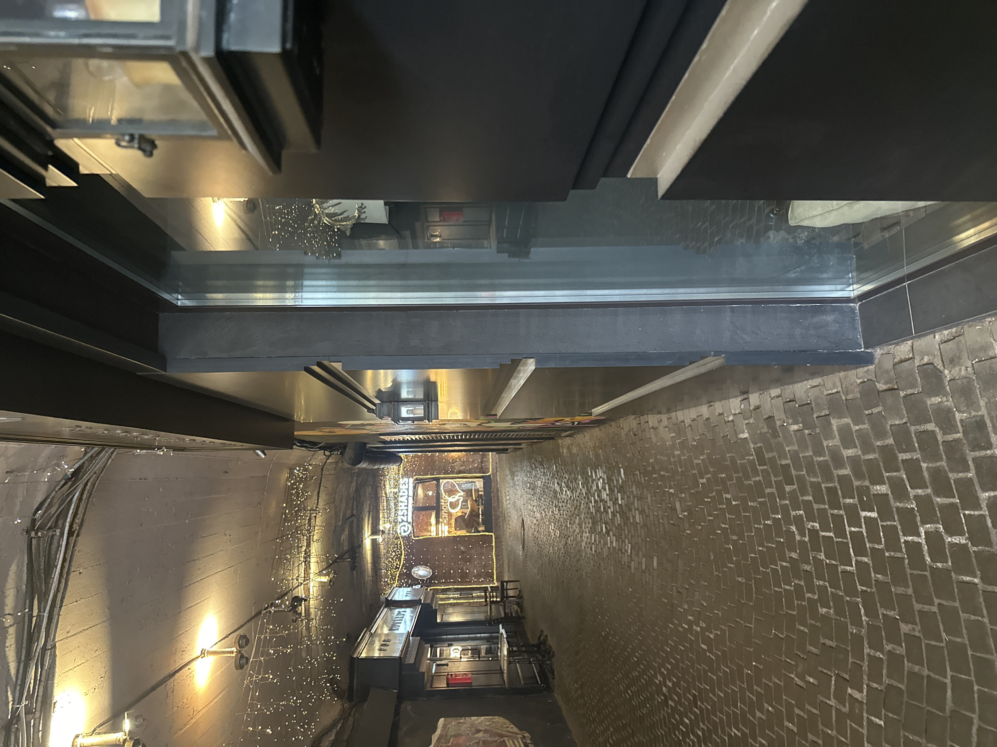 The image shows a narrow street or alleyway in Moscow, Russia, as viewed from a high vantage point, possibly through a window or from a balcony. The alleyway is paved with dark, rectangular stones arranged in a running bond pattern.
The left side of the alleyway has a light-colored wall with exposed electrical conduits and lights, some decorated with strings of small, bright lights. A business with the name "2 SHADES" is visible further down the alleyway, also adorned with decorative lights. Further down a building with signs with russian letters.
The overhead view includes architectural details of the vantage point, featuring dark-colored window frames and possibly part of a ceiling or balcony structure. The lighting in the alleyway suggests a mix of natural and artificial light sources.