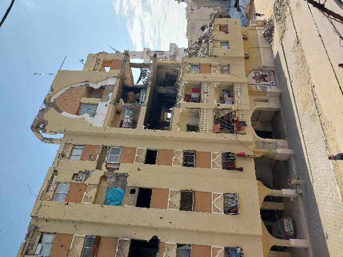 The image displays a multi-story residential building in Khan Yunis, Palestinian Territory, bearing extensive damage indicative of conflict. The structure, rendered in light beige with brown decorative accents, stands beneath a clear blue daytime sky.
The upper floors, particularly towards the left side, show severe structural collapse, with large sections of walls and ceilings completely missing, exposing internal rooms and twisted rebar. A significant area of black charring and fire damage is prominent in the mid-upper section of the building, suggesting an intense blaze. Many windows throughout the building are broken or entirely absent.
Despite the widespread destruction, evidence of continued life is visible. On the right side of the building, clotheslines with laundry are hanging from balconies and window openings on several floors, implying recent or ongoing human presence. Air conditioning units are also mounted on the exterior walls.
At street level, the building features arched openings, with a white car partially visible under one. The adjacent paved street and sidewalk are littered with debris. Several individuals are present on the street, some walking, others standing, in the vicinity of the damaged building. On an arched wall near the ground, there is graffiti depicting a red tractor. The scene conveys a strong impression of devastation, illustrating the profound impact of conflict on urban infrastructure and daily life.