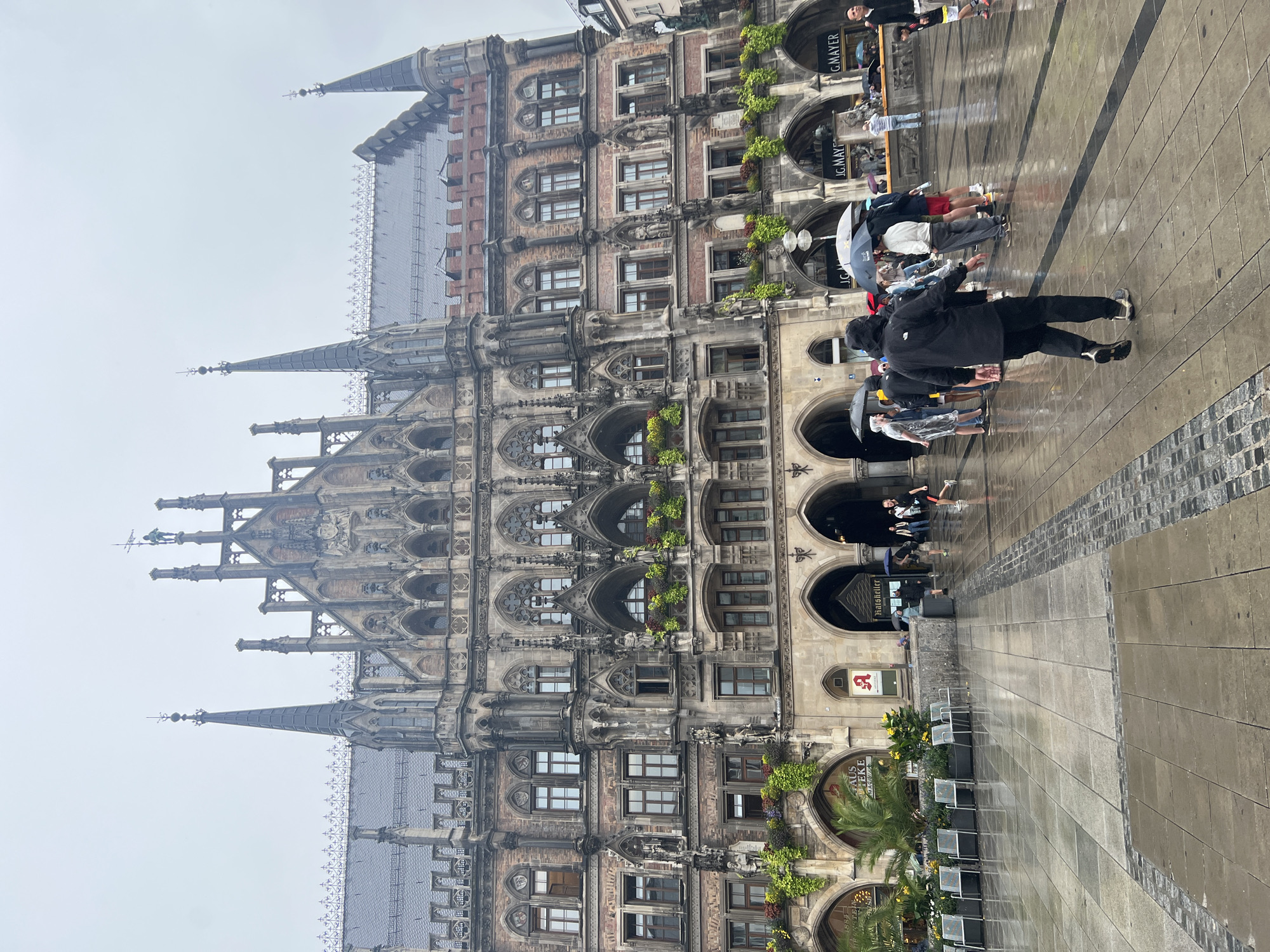 The image depicts a frontal view of the New Town Hall (Neues Rathaus) in Munich, Germany, on a rainy day. The architecture is Gothic Revival, characterized by intricate stonework, pointed arches, spires, and numerous windows. Several people are present in the square in front of the building, many holding umbrellas due to the rain. The ground is wet and reflective, indicating recent or ongoing precipitation. Some individuals are walking, while others appear to be standing in groups. The scene captures a typical public space in a European city during inclement weather, with the grand facade of the Rathaus serving as a prominent backdrop.