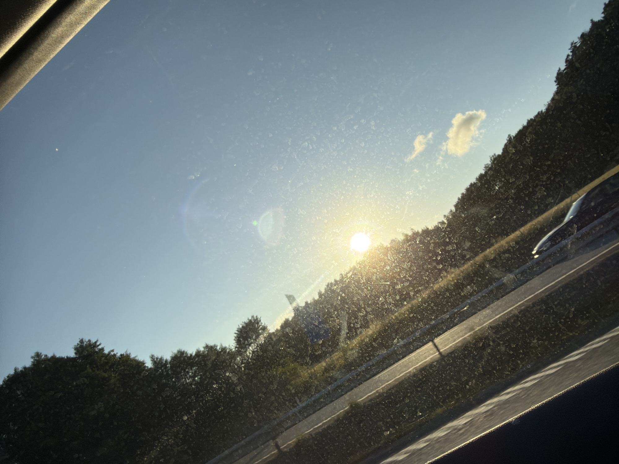The image captures a sunlit scene, possibly from inside a vehicle, looking out through a window with visible dust or water spots. The perspective is angled, with the foreground dominated by a dark, possibly upholstered surface on the right and a diagonal line suggestive of a window frame or dashboard. A road, marked by white lines, runs diagonally across the lower left portion of the frame, bordered by dense, dark green foliage that appears to be trees or bushes. In the upper right, a clear blue sky is visible, with a few small white clouds and a bright, blurred sun. The sun is low in the sky, casting a warm glow and creating lens flare. There is a hint of a vehicle's reflection or interior in the very bottom center of the image. The scene appears to be a roadside view, potentially along a highway, outside of Stenungsund, Sweden, given the typical landscaping and road infrastructure found in such areas.