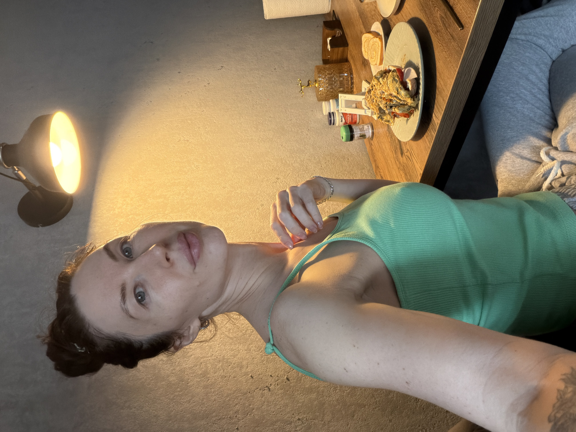 A woman is taking a selfie at a table. She has her hair up in a bun and is wearing a green tank top. The table is brown and has a plate of food on it, as well as some pills and spices. There is a lamp on the left side of the image. The location is in Perm, Russia.