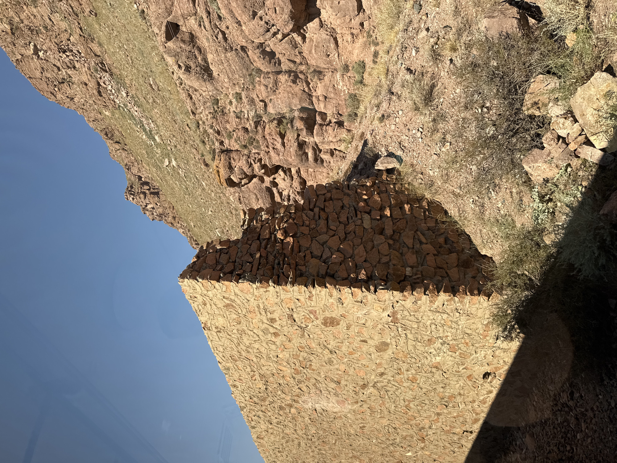This image depicts a stone wall in a rocky, mountainous environment, under a clear blue sky. The wall is constructed from rough, irregularly shaped stones of varying sizes and shades of brown and orange, with visible mortar between them. The wall appears to be part of a ruin or an old structure, as it is not perfectly intact and shows signs of age.
To the right of the wall, the terrain rises sharply into a steep, rocky mountainside. This mountainside is characterized by exposed red rock formations, patches of dry grass, and sparse vegetation. The sunlight casts a distinct shadow from the wall onto the ground, indicating a bright, sunny day. The foreground shows a mixture of loose stones, gravel, and low-lying, dry brush.
Without further context or identifying features within the image, it is not possible to definitively pinpoint the location in Krasnyy Most, Kyrgyzstan. The scene suggests a remote, undeveloped area, likely in a mountainous region where such stone structures might be found, possibly remnants of historical settlements or fortifications. There are no people, animals, or any discernible activities taking place. The image is a static, environmental portrait of the wall and its natural surroundings.