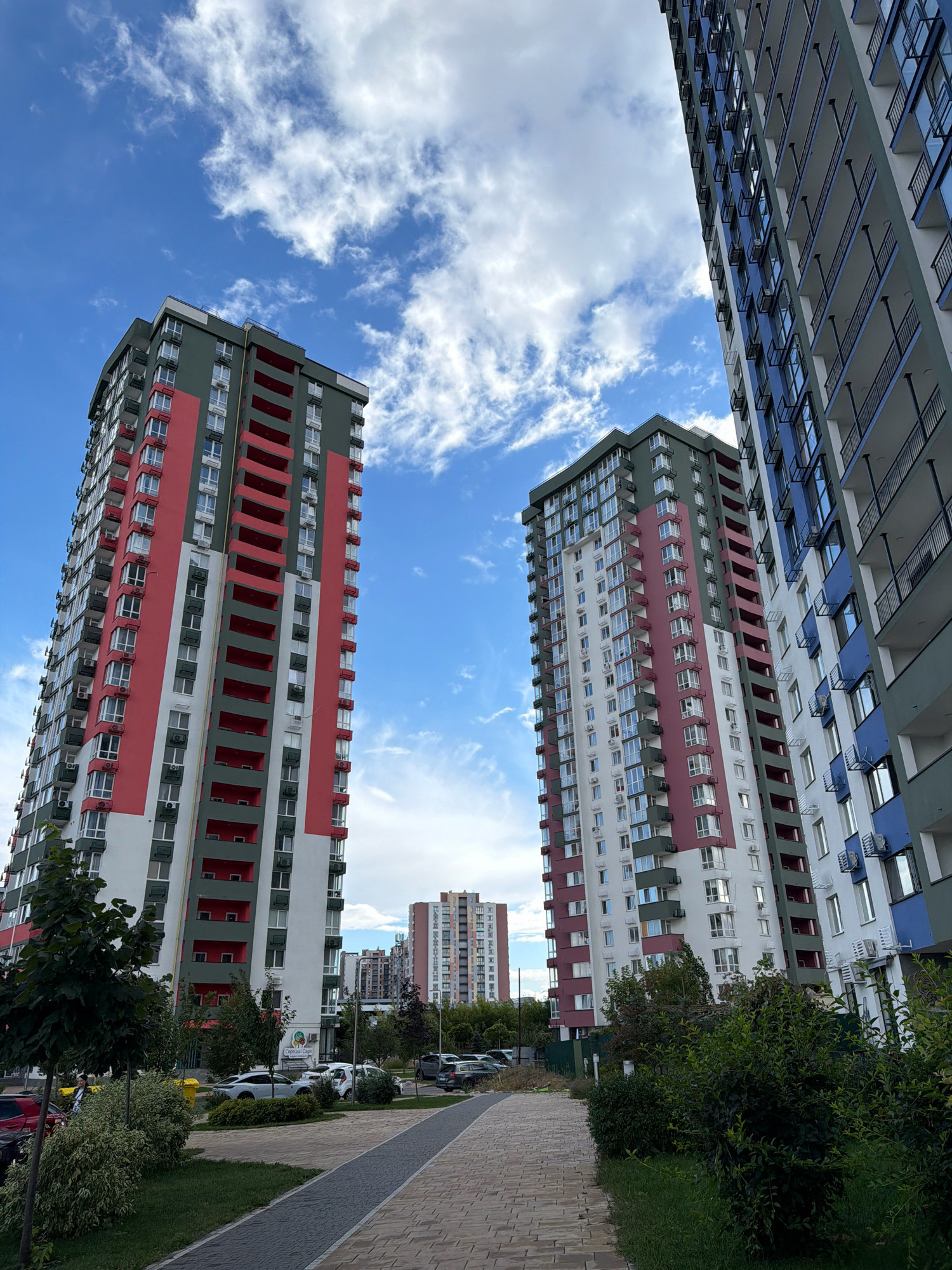 In Kyiv, Ukraine, this image captures a contemporary residential area dominated by several high-rise apartment buildings. The sky is a vibrant blue, dotted with scattered white clouds. In the foreground, a paved walkway with a decorative pattern leads through a green space with manicured trees and bushes. Cars are parked along the side of the walkway.
The most prominent structures are three tall apartment buildings. The building on the left is primarily colored in sections of dark green, white, and bright red, with balconies accentuating its height. To its right, a similar building displays a color scheme of dark green, white, and a deep pinkish-red. Further to the right, another building is visible, predominantly white with accents of dark blue and grey, also featuring numerous balconies. In the distance, more apartment buildings of varying heights and designs are visible, indicating a densely populated urban environment. The architecture suggests modern construction, with numerous windows and external air conditioning units visible on the facades of the buildings.