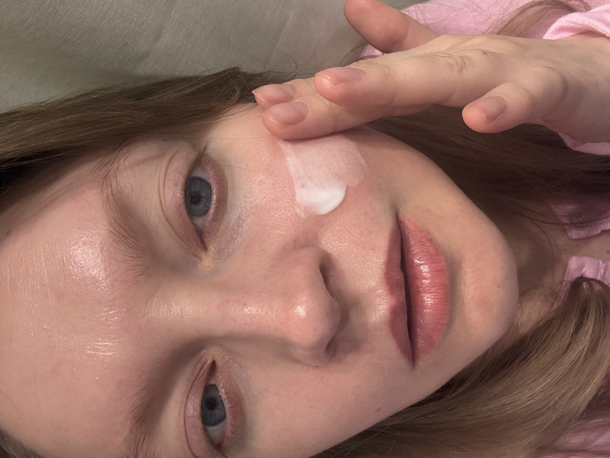 A close-up, tilted portrait shows a fair-skinned woman applying a white cream to her face. Her right hand touches her right cheek, spreading the cream. She has blue eyes, pink lips, and light brown hair. She is wearing a pink shirt. The background is a light gray/beige color. The image does not contain enough information to identify the location as Izhevsk, Russia.