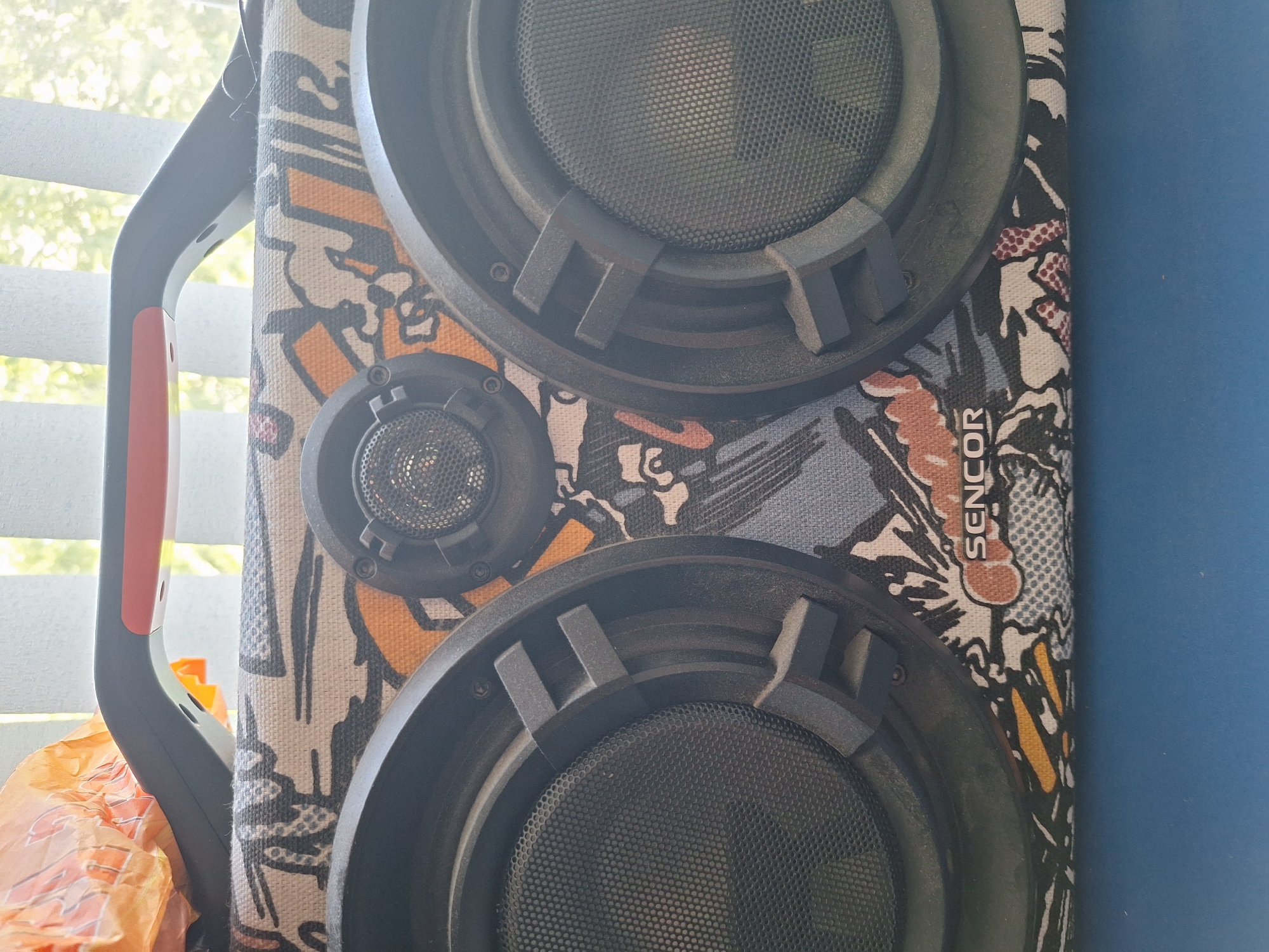 A close-up shot shows a Sencor brand portable speaker. The speaker is covered in a colorful, comic-book-style graphic print with black, white, orange, and blue elements. It has a black handle with orange accents on top, and two large black speaker grilles on the front. There is also a smaller speaker or tweeter located centrally above the two main speaker grilles. The speaker sits on a blue surface. In the background, a window with vertical blinds is visible, with blurred green foliage outside, suggesting an indoor setting, possibly a room with a view of trees or a garden. The image does not contain enough information to determine its location within the city of Varna, Bulgaria.