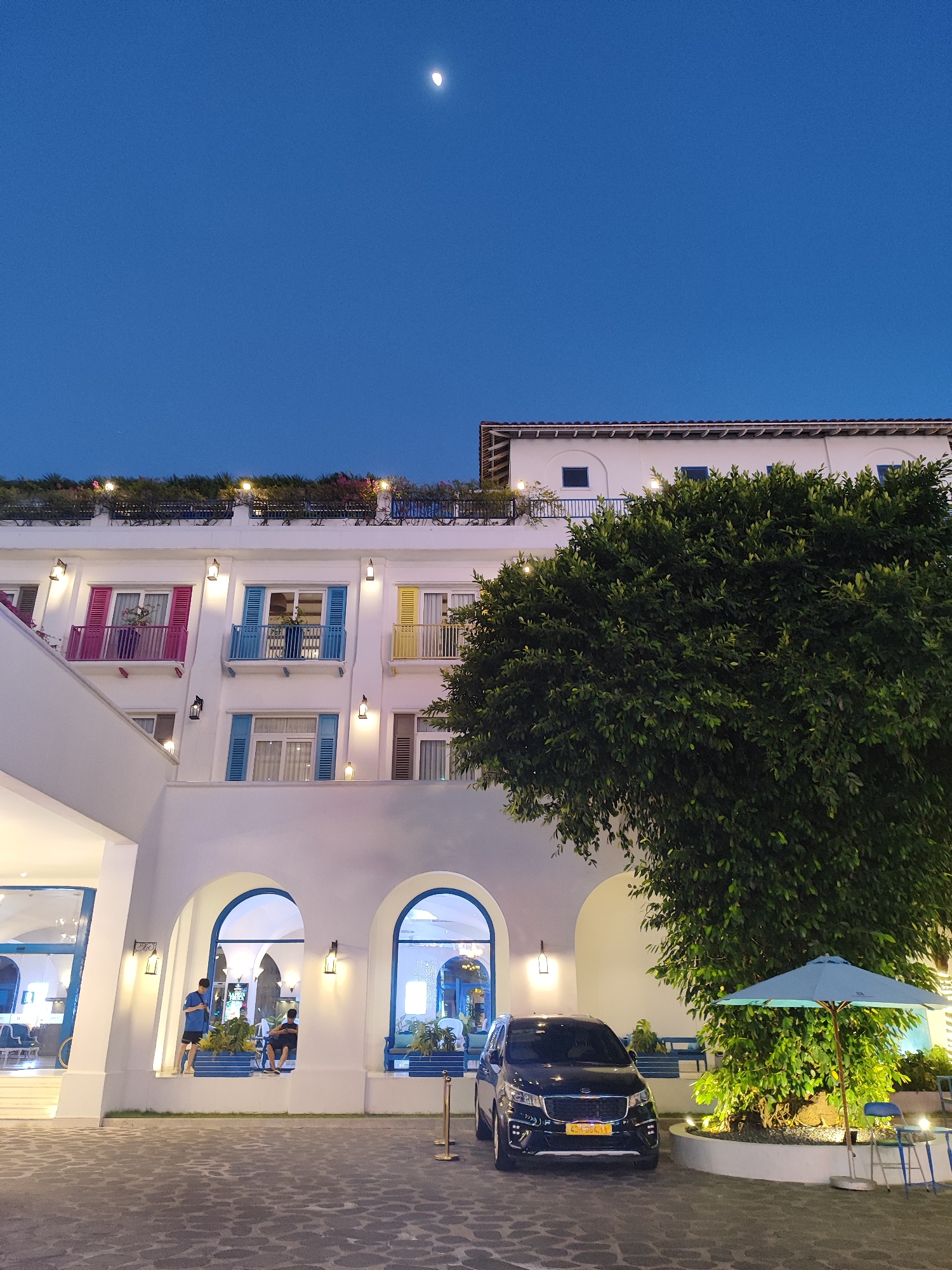 The image depicts the exterior of a brightly colored building, possibly a hotel or resort, with a distinctly Mediterranean architectural style, featuring white walls and colorful shutters on the windows in shades of pink, blue, and yellow. The scene is set at dusk, as indicated by the deep blue sky and the presence of a crescent moon. A lush, full-grown tree with dark green foliage dominates the right side of the frame, partially obscuring a portion of the building and providing a stark contrast to the white structure.
In the foreground, a dark-colored minivan is parked on a cobblestone-like pavement. Next to the tree, a light blue umbrella shades an outdoor seating area with a small table and chairs. The building's entrance features arched doorways leading into illuminated interiors, where a few people can be seen. One person stands near an archway, appearing to be an employee or resident, while others are seated inside what looks like a cafe or lounge area. Small outdoor lights are strategically placed along the building's facade, adding to the ambiance.
The location is identified as Đà Nẵng, Vietnam. The overall atmosphere suggests a relaxed evening setting, perhaps a tourist destination or a residential complex, with a blend of architectural beauty and natural elements. The presence of the moon and the darkening sky point to the transition from day to night. The minivan has a license plate with text, which appears to be "43H-556.91".